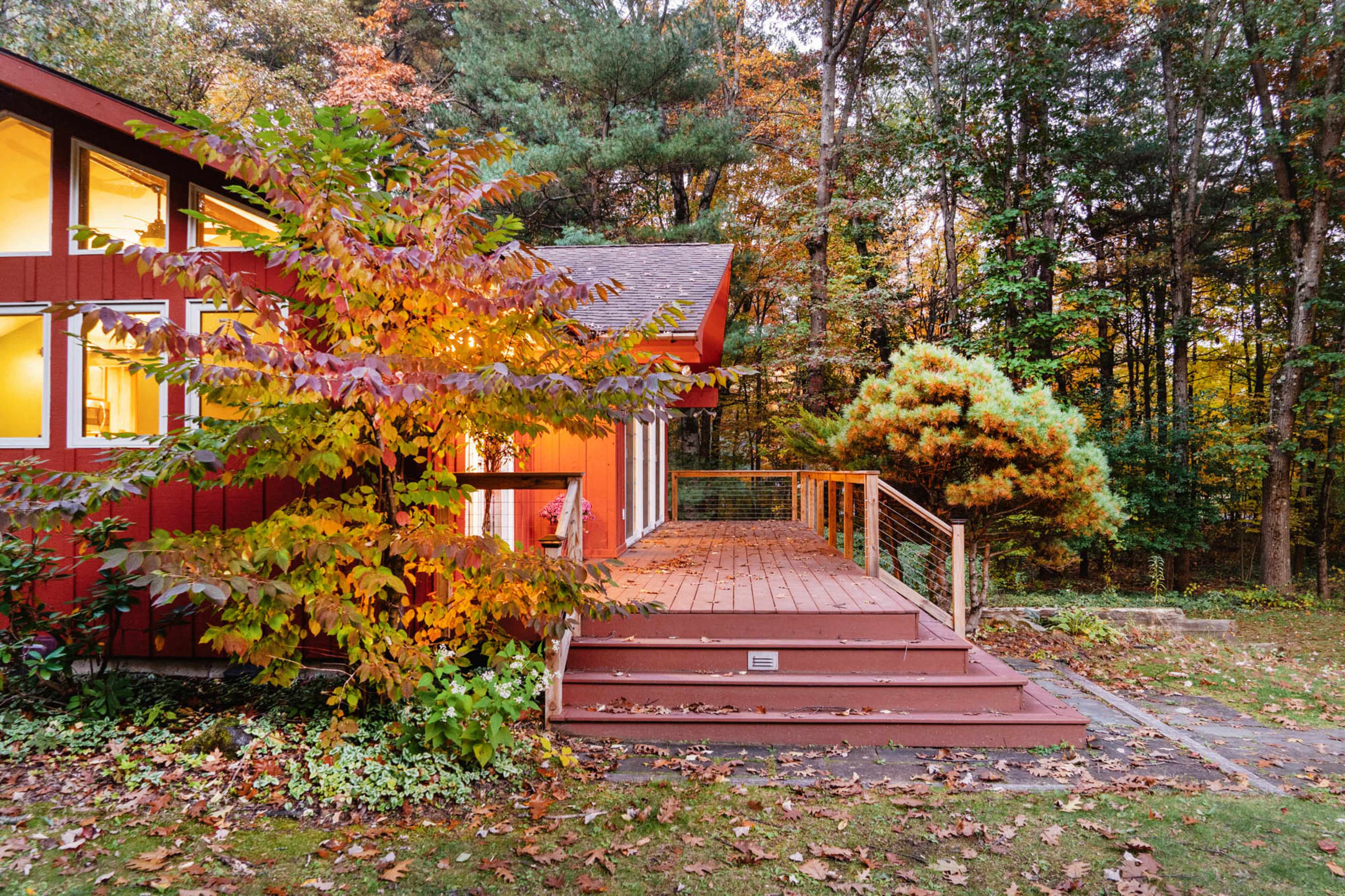 A red house with large windows has a wooden deck surrounded by colorful autumn foliage.