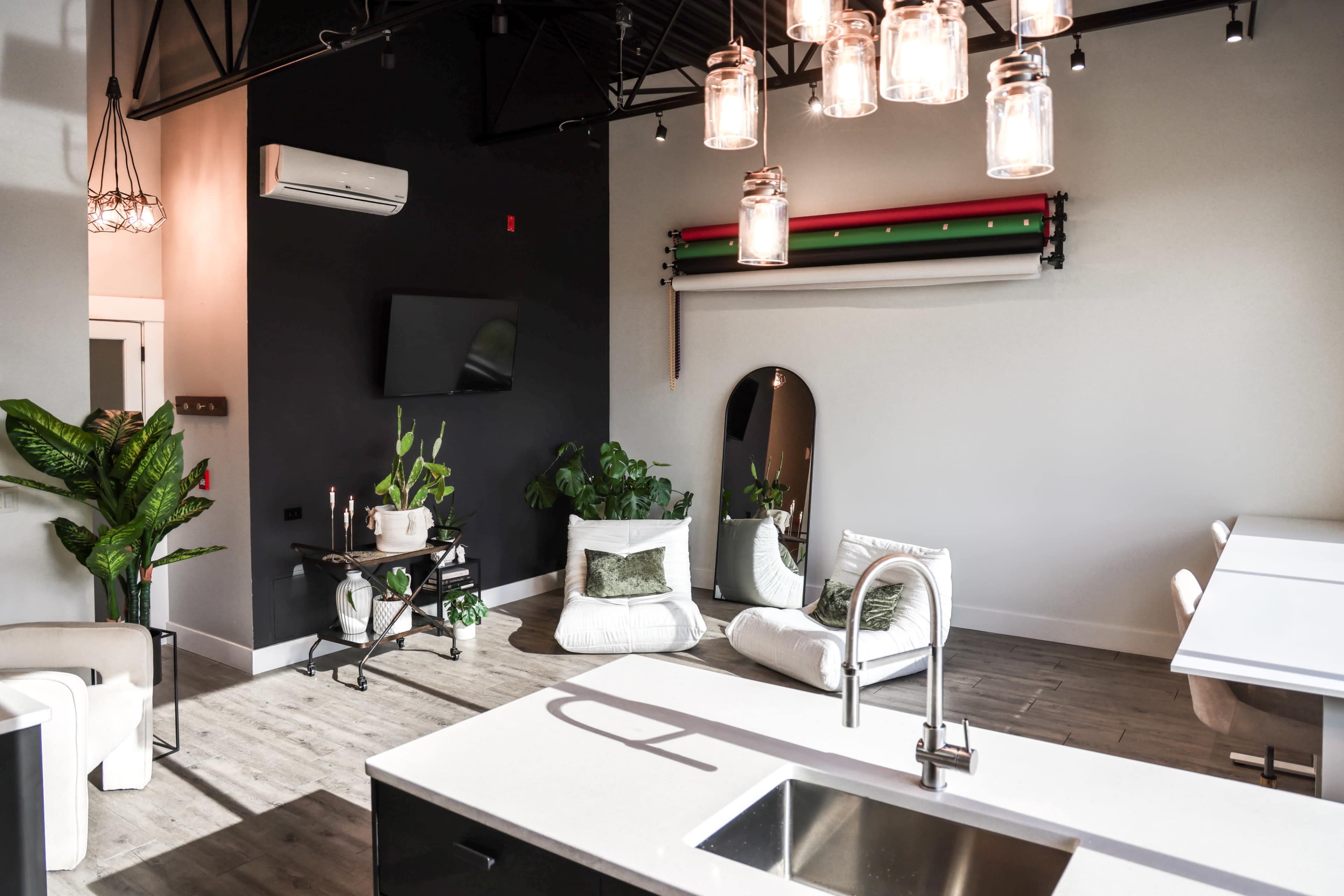 The image shows a modern interior space featuring a kitchen island, comfortable seating, and greenery, illuminated by pendant lights.