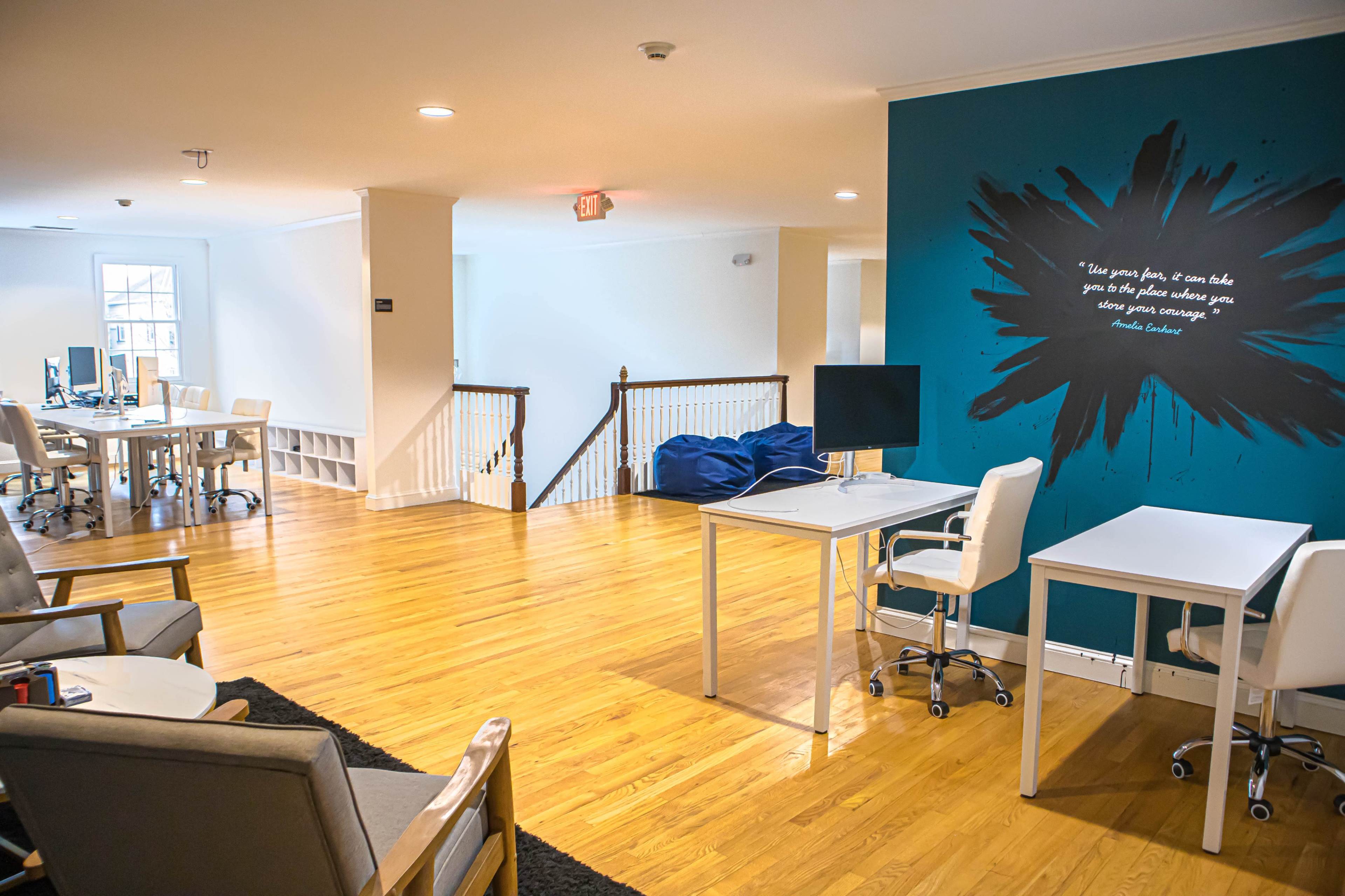 The image shows a modern office space featuring wooden floors, white tables, and a blue accent wall with a quote.