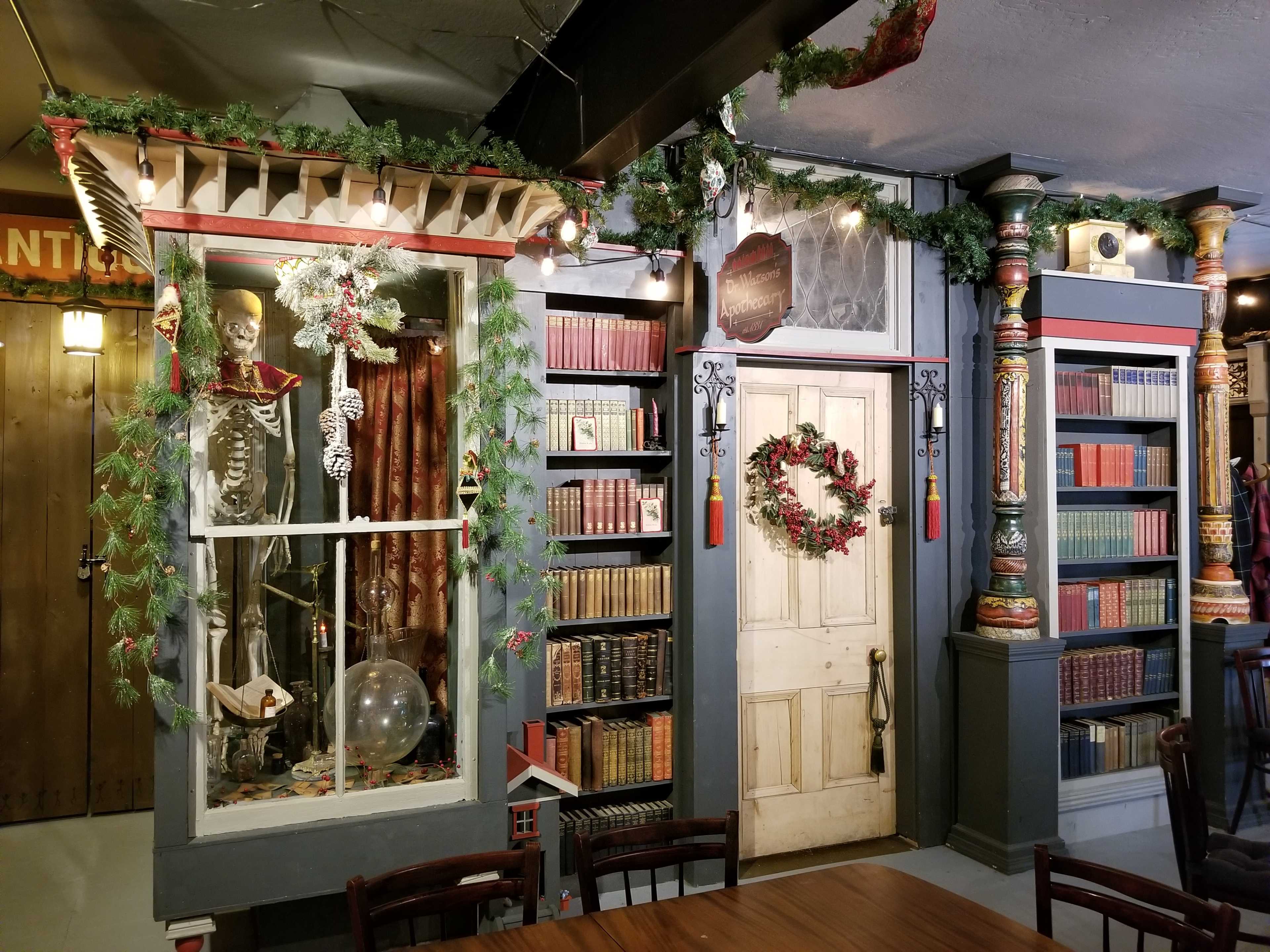 A corner of a room featuring a skeleton in a display case, surrounded by bookshelves filled with books and festive decorations, including garlands and a wreath on the door.