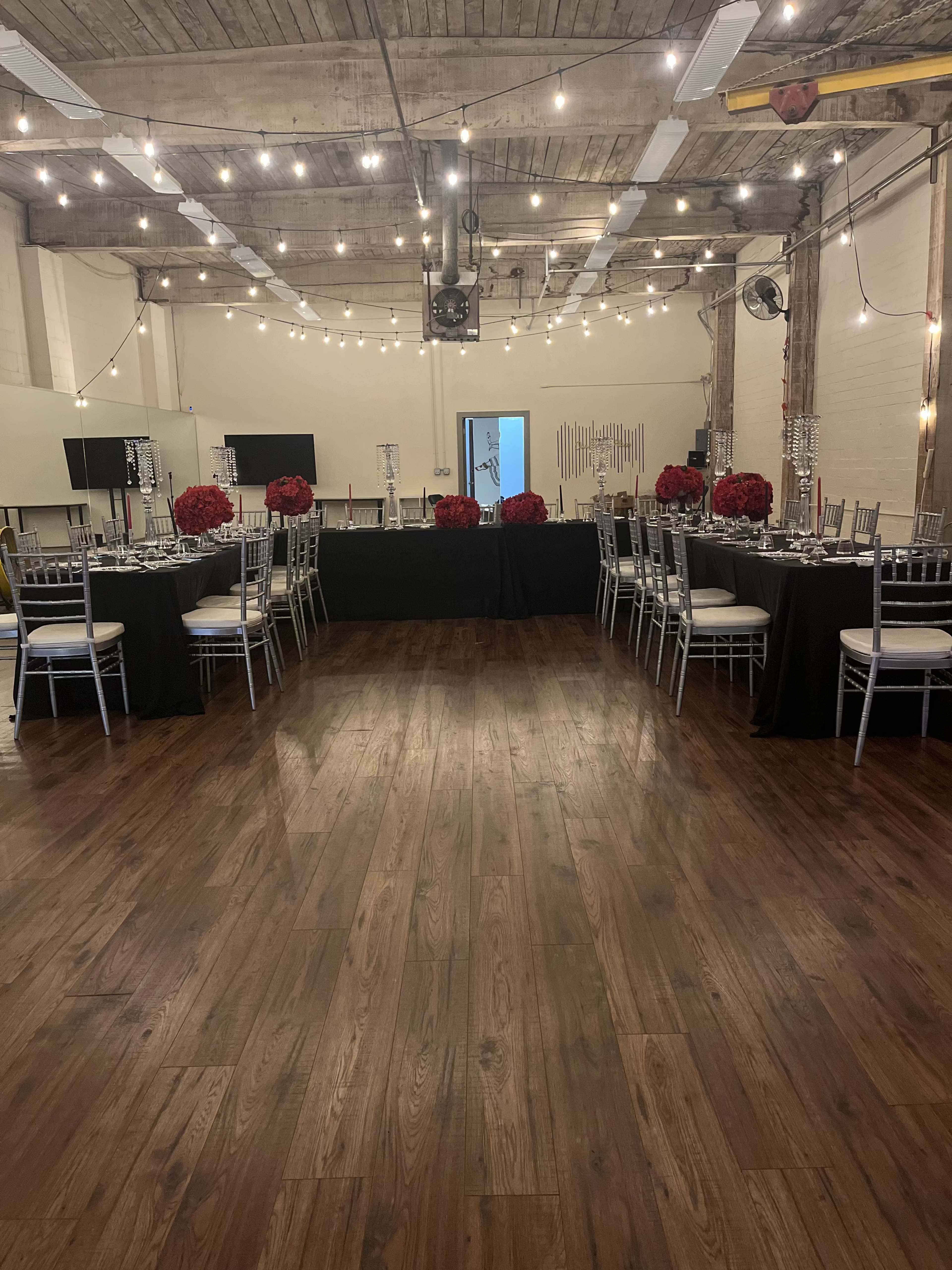 A spacious event venue features tables set with dark tablecloths and elegant centerpieces, surrounded by wooden flooring and string lights overhead.