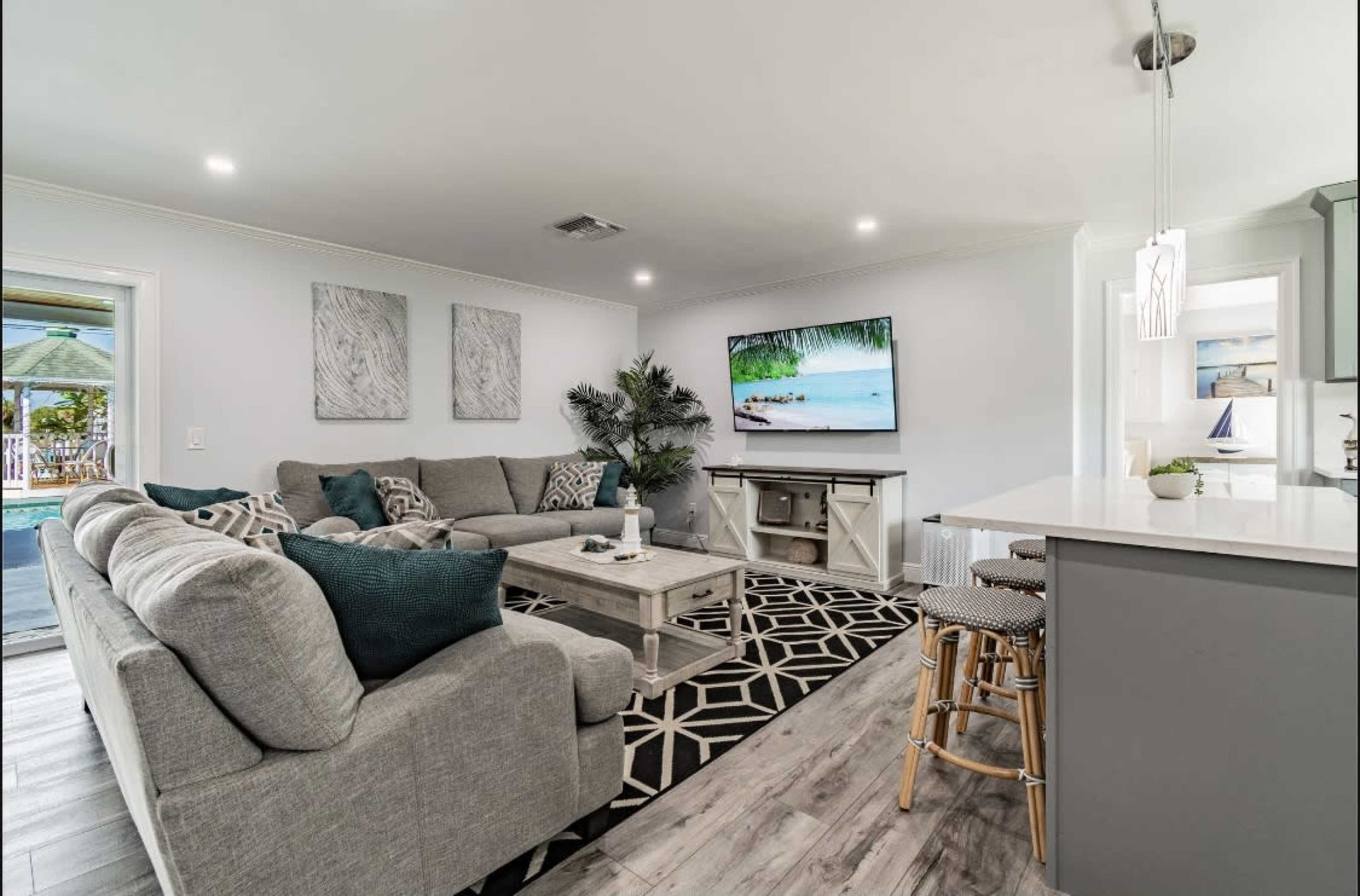 A modern living room features a large gray sectional sofa, a coffee table, and a flat-screen TV displaying a scenic beach image, with a kitchen area visible in the background.
