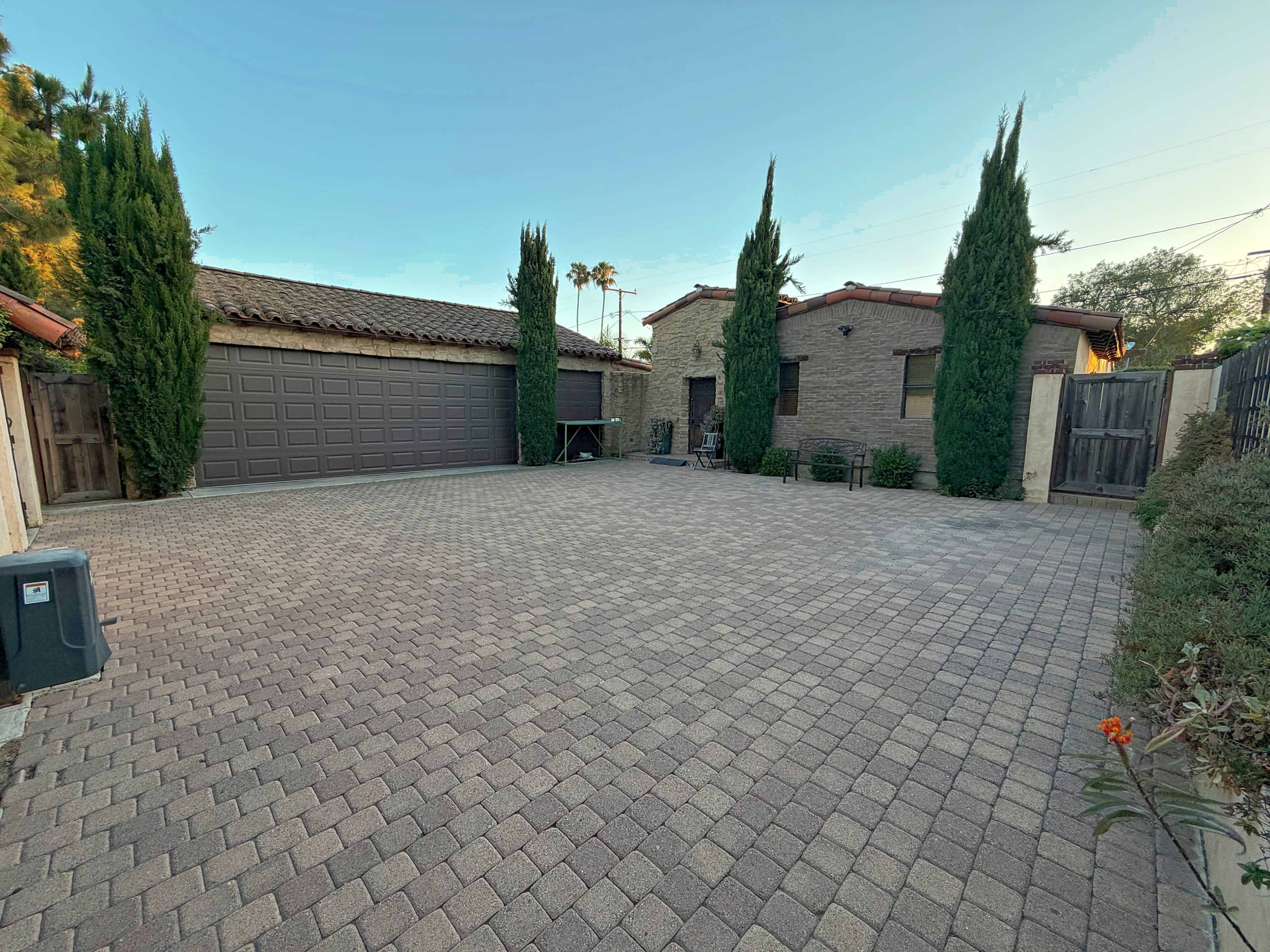 The image shows a spacious paved outdoor area with two buildings on either side and several tall, narrow trees in the background.