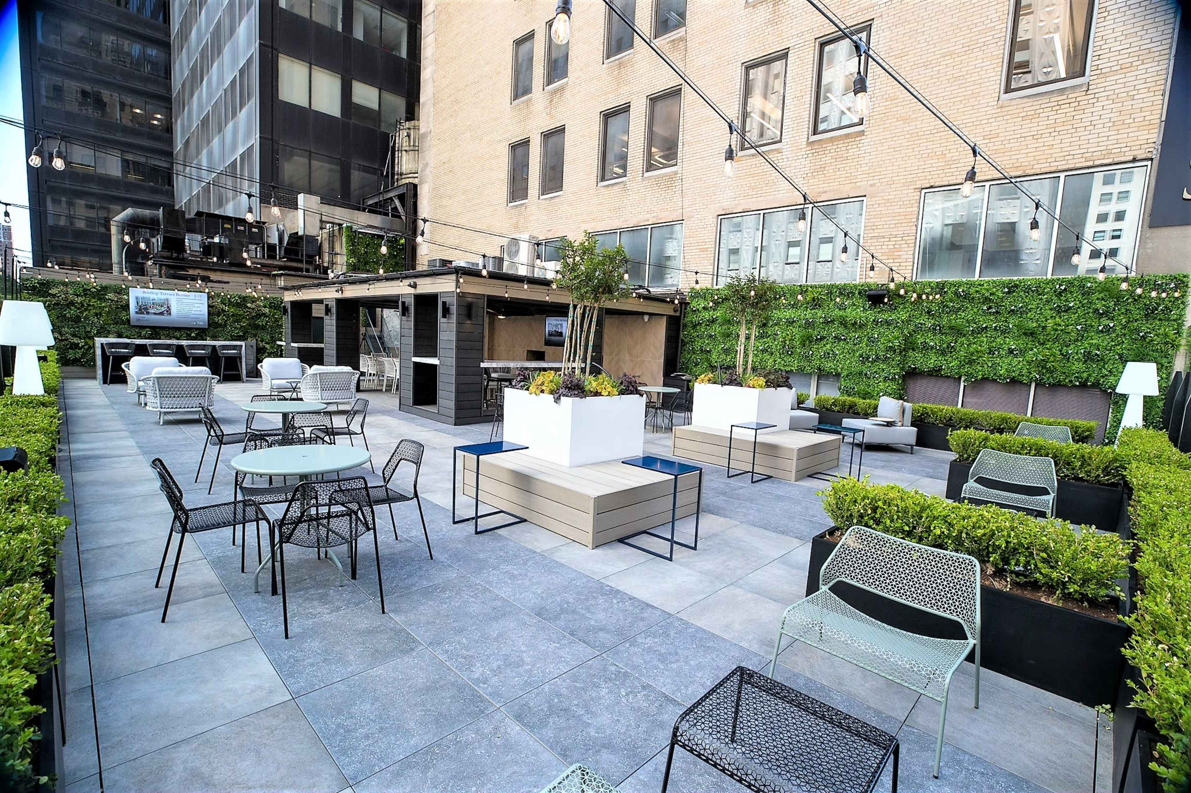 The image shows a modern rooftop patio area with tables and chairs, surrounded by greenery and partially enclosed by buildings.