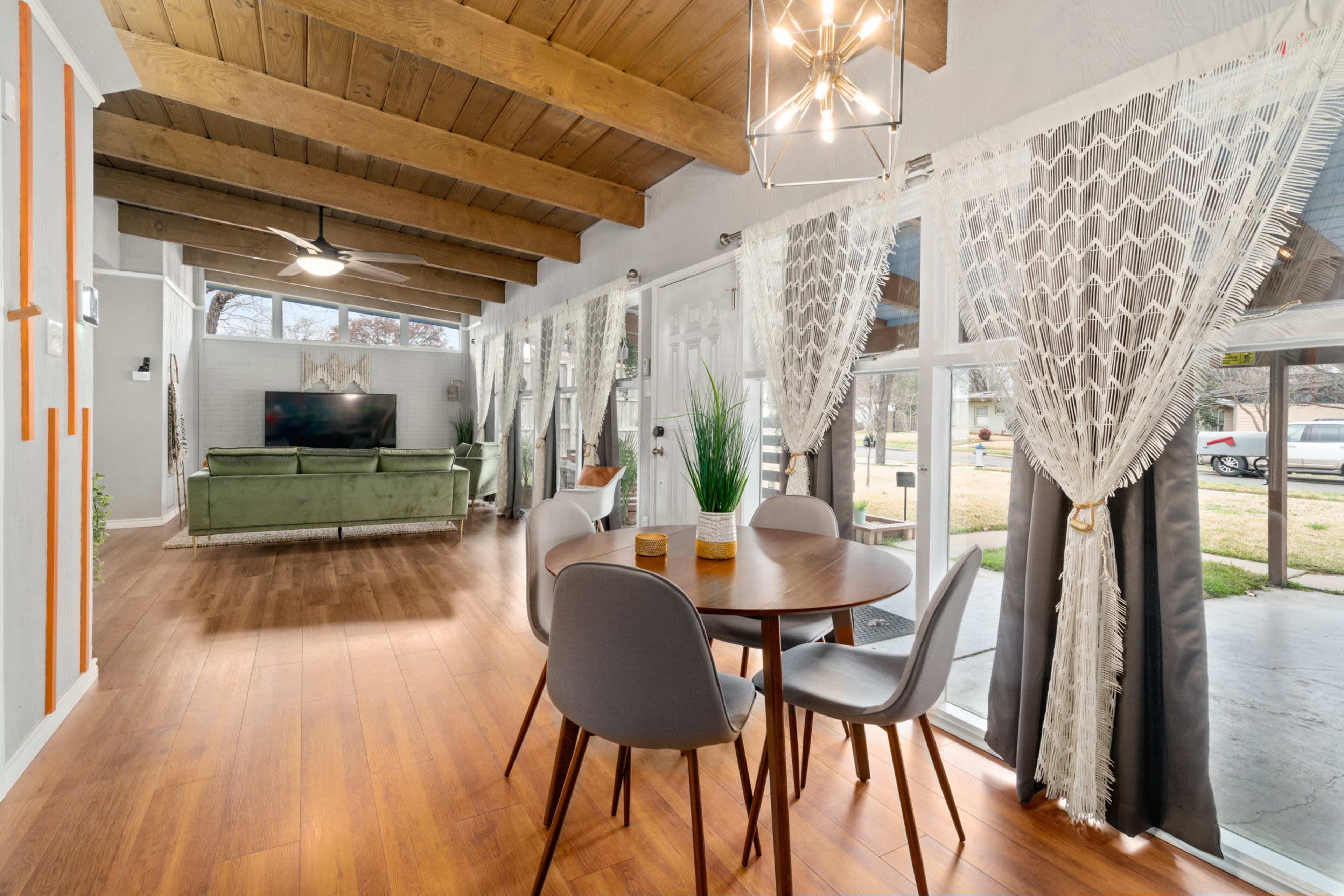 A dining area features a round table surrounded by chairs, with large windows showcasing a living space adorned with a green sofa and a television.