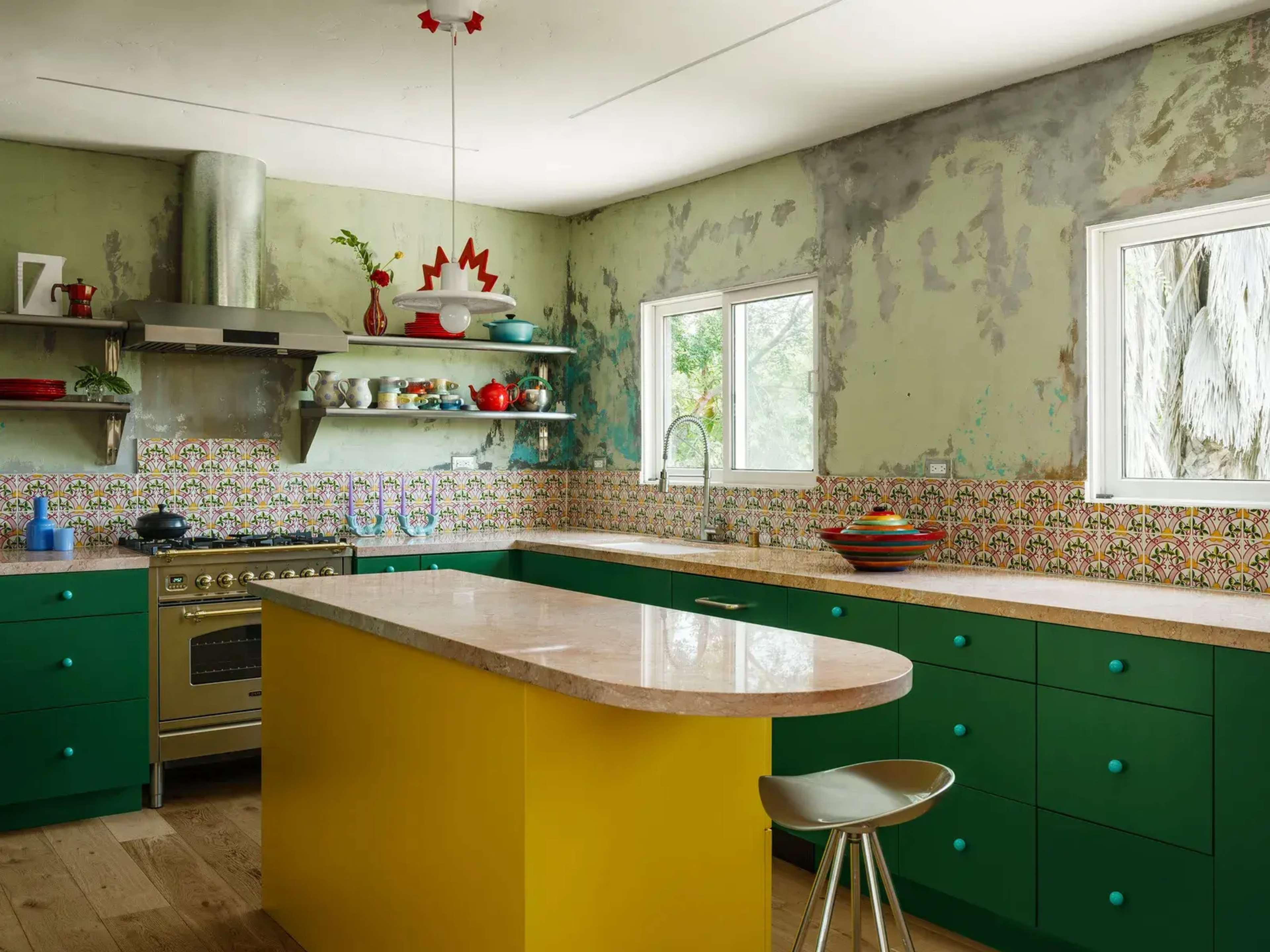 The kitchen features a yellow island with green cabinet bases, a stainless steel oven, and colorful dishware on the open shelves.