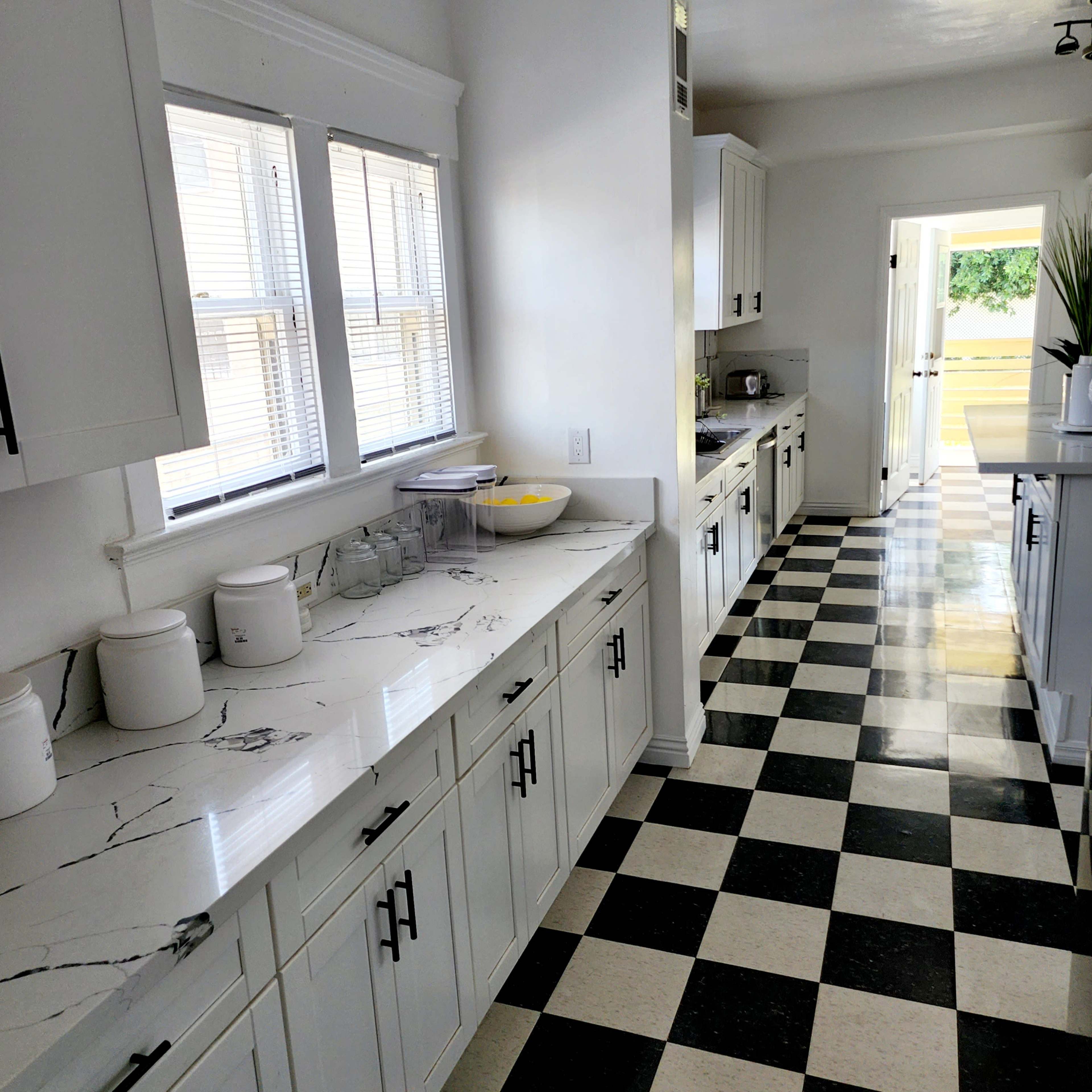 A bright kitchen features white cabinets, a marble countertop, and a black and white checkered floor.