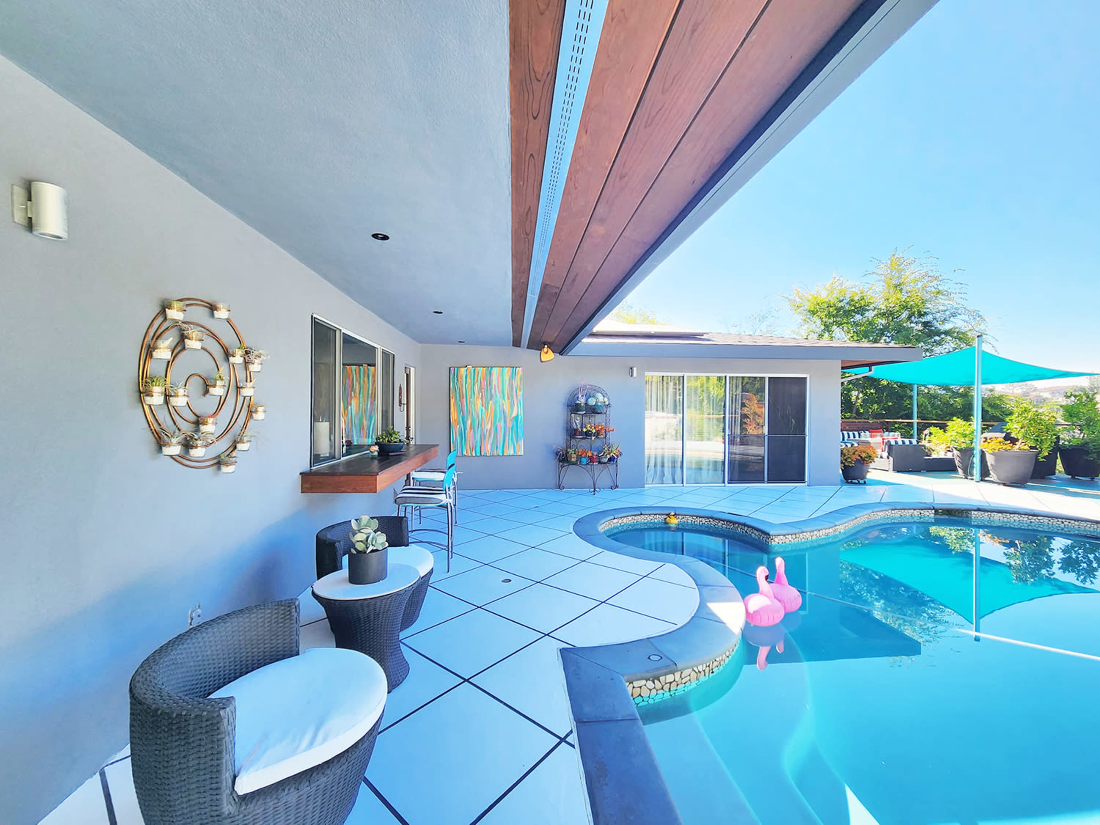 The image shows a modern patio with a swimming pool, two chairs, and colorful artwork on the walls under a clear blue sky.