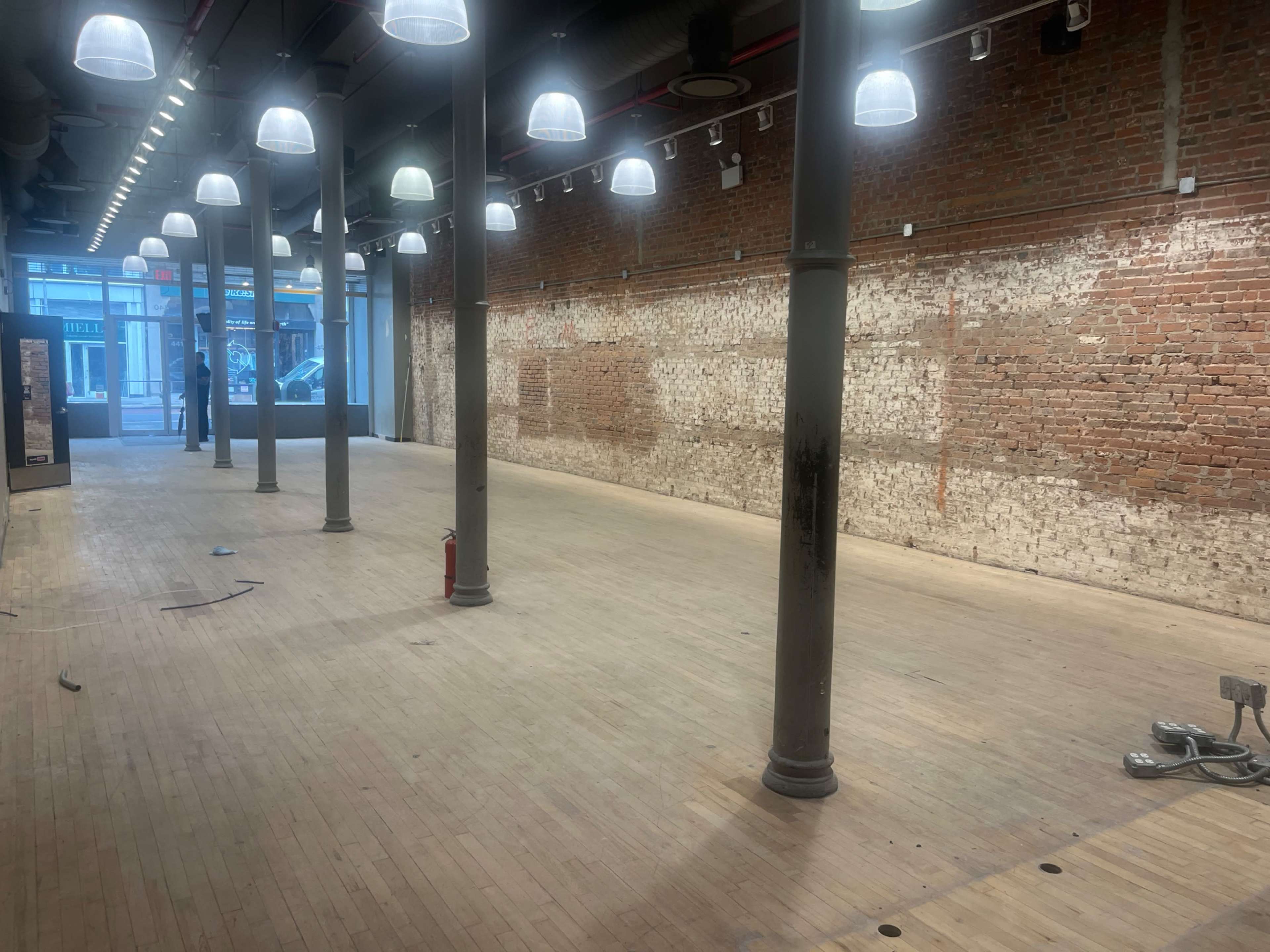 The image shows an empty interior space with wooden flooring, exposed brick walls, tall columns, and overhead lighting.
