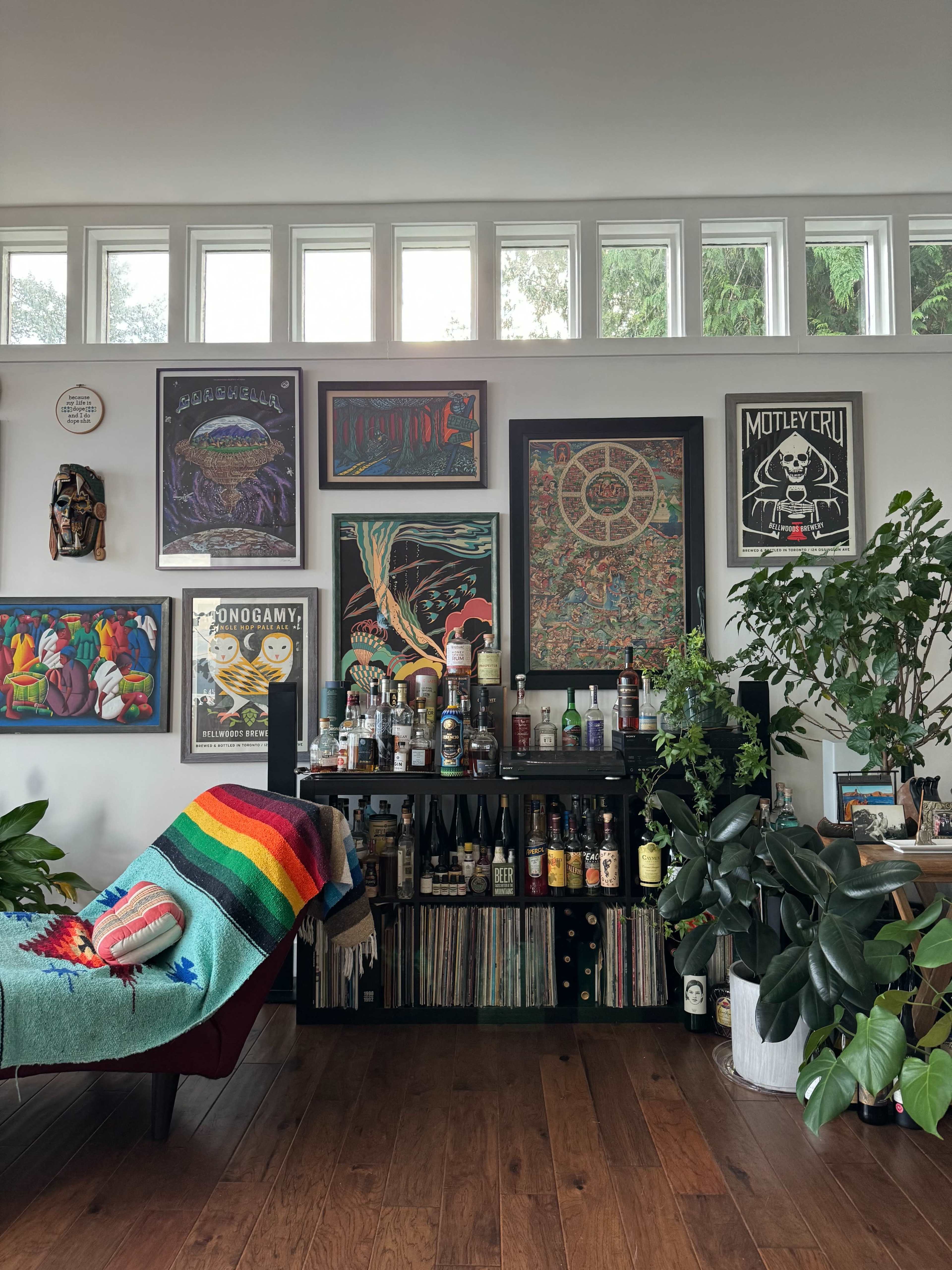 A colorful wall adorned with various framed posters and artwork above a shelf filled with bottles and vinyl records, accompanied by a couch draped with a rainbow-striped blanket and several green plants.