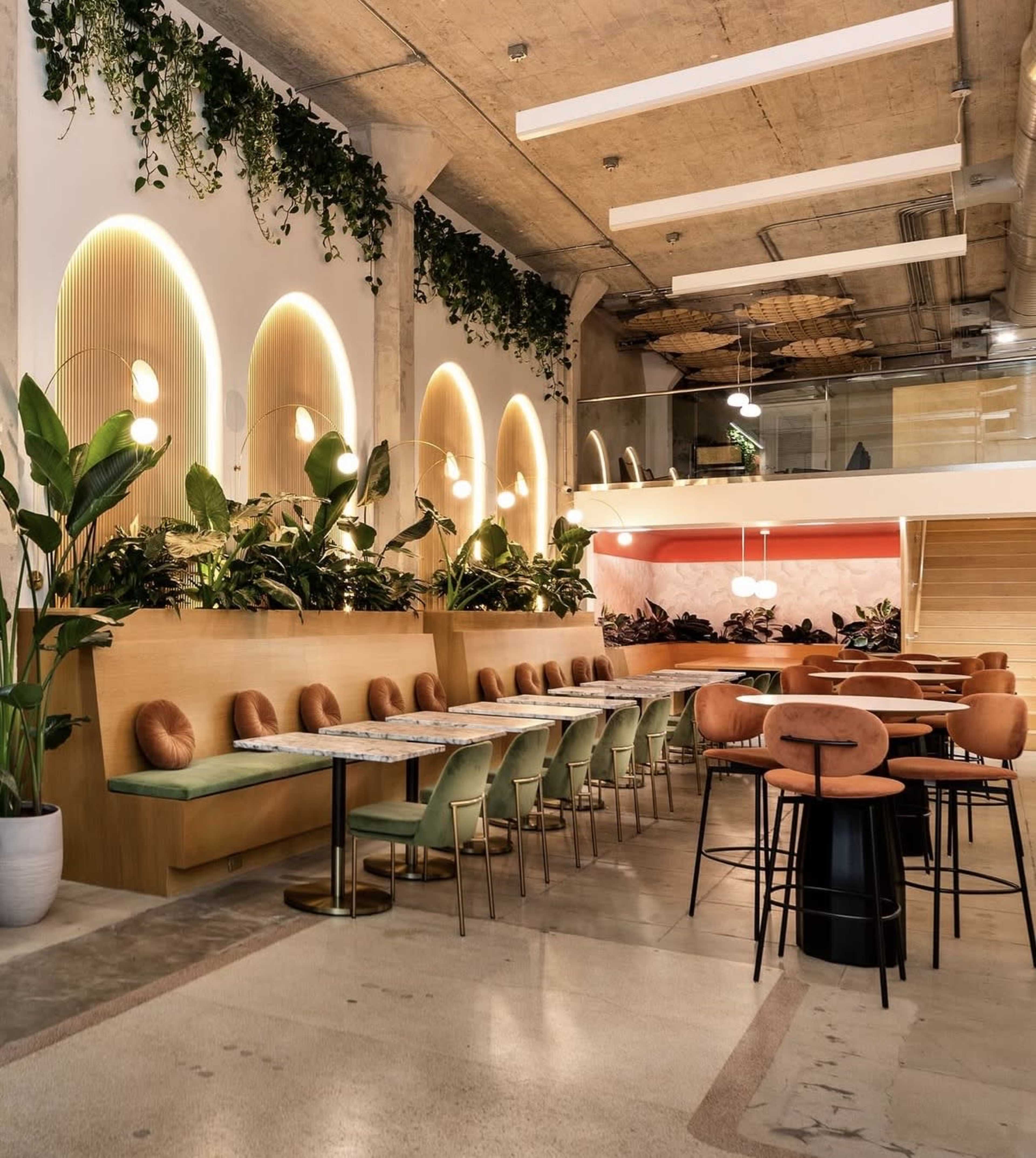 The image shows a modern restaurant interior featuring a long communal table surrounded by green chairs, with lush plants and soft lighting adorning the walls.