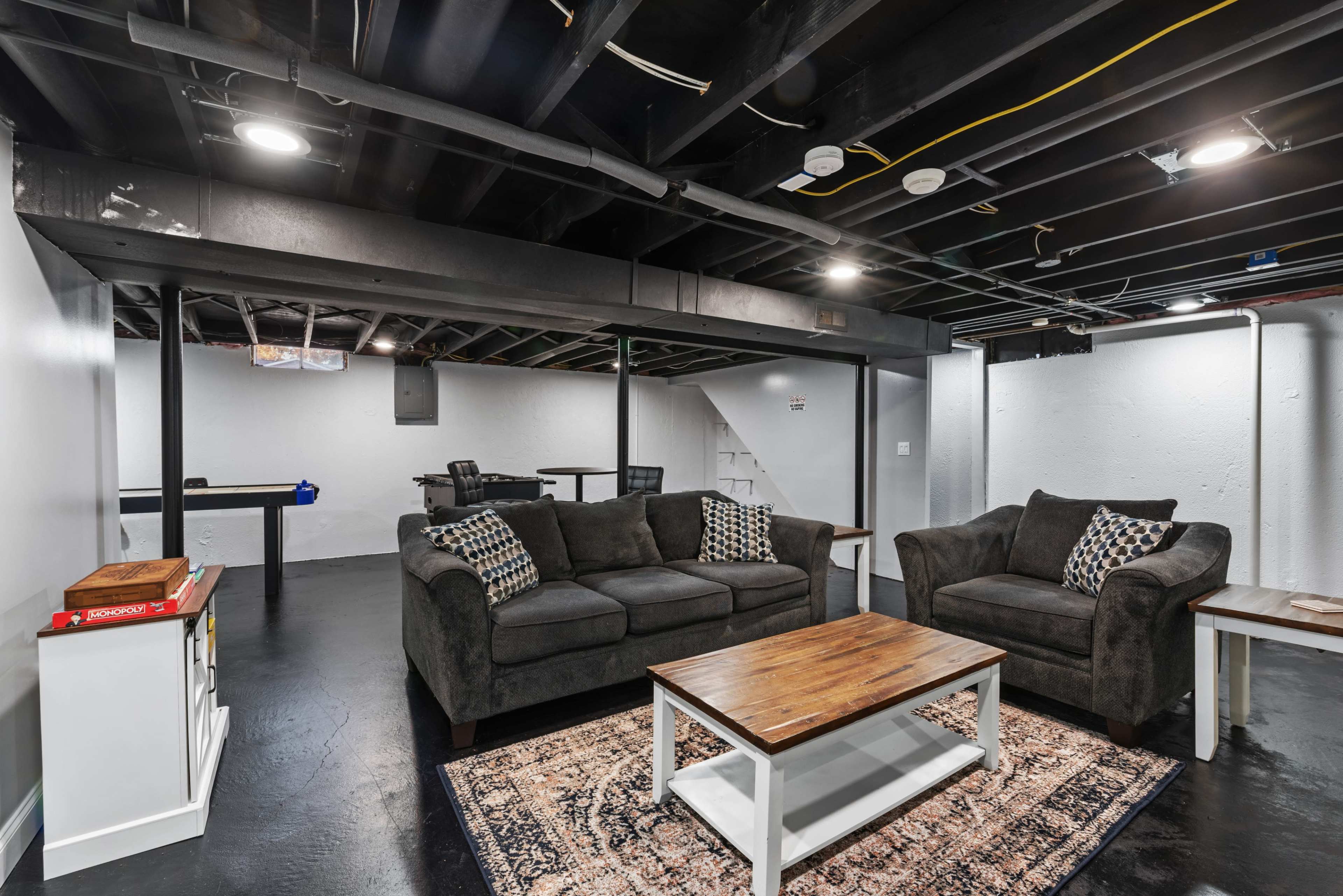 A finished basement with black ceilings, a coffee table, two dark sofas with pillows, and a small dining area.