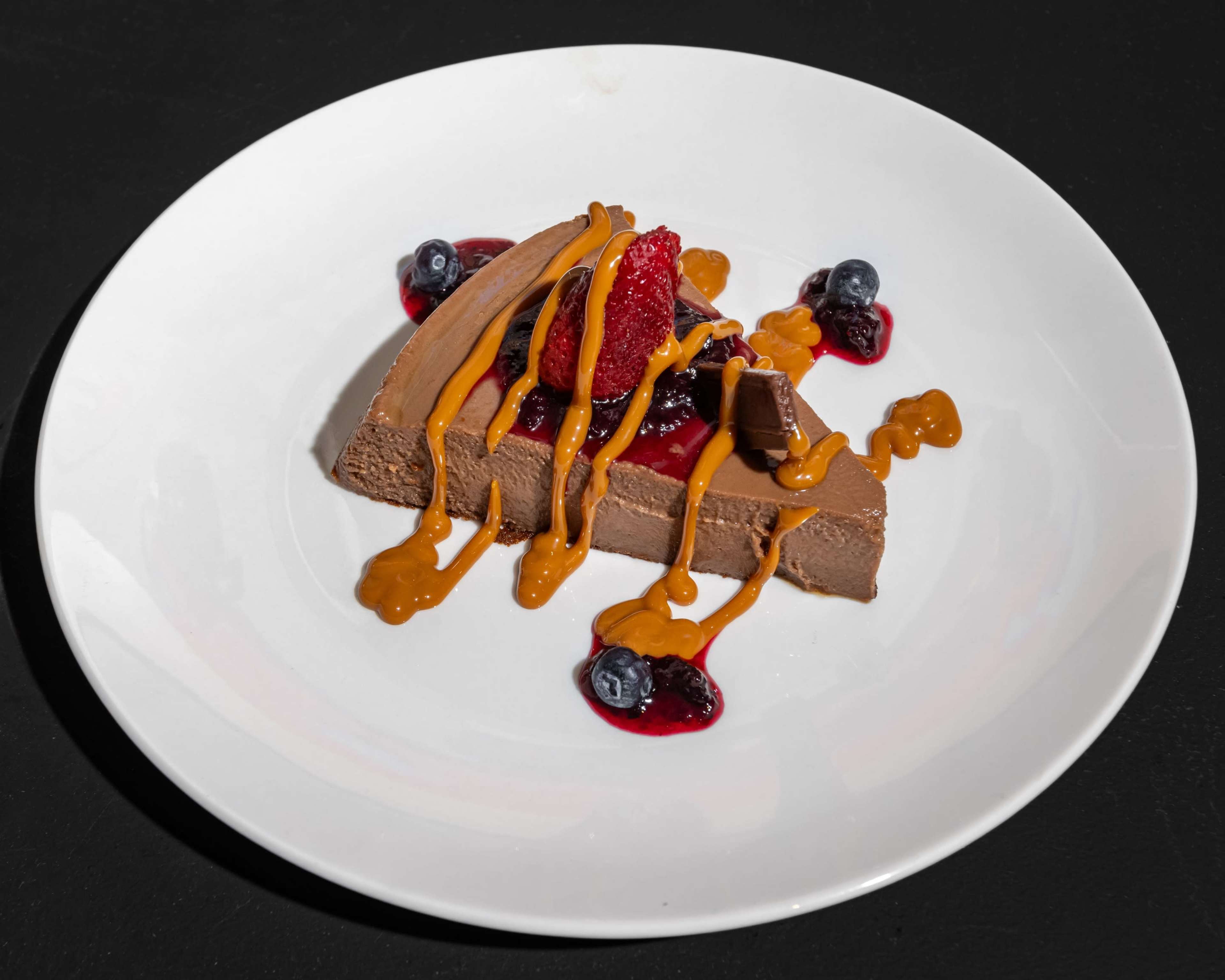 A slice of chocolate dessert is topped with strawberries, blueberries, and a drizzle of sauce on a white plate.