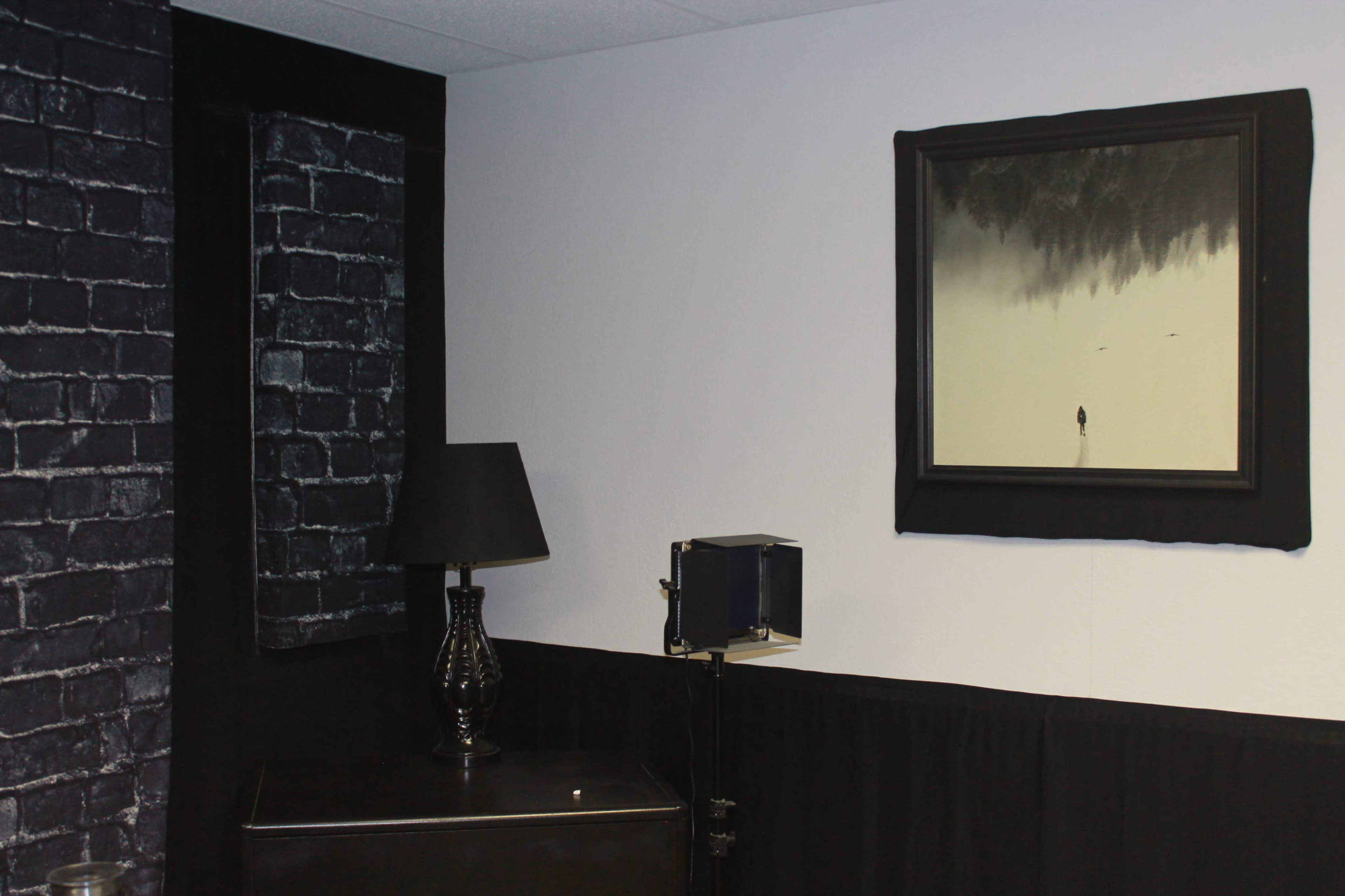 A room with a black brick wall panel, a lamp on a black dresser, and a framed painting depicting a solitary figure in a foggy landscape.