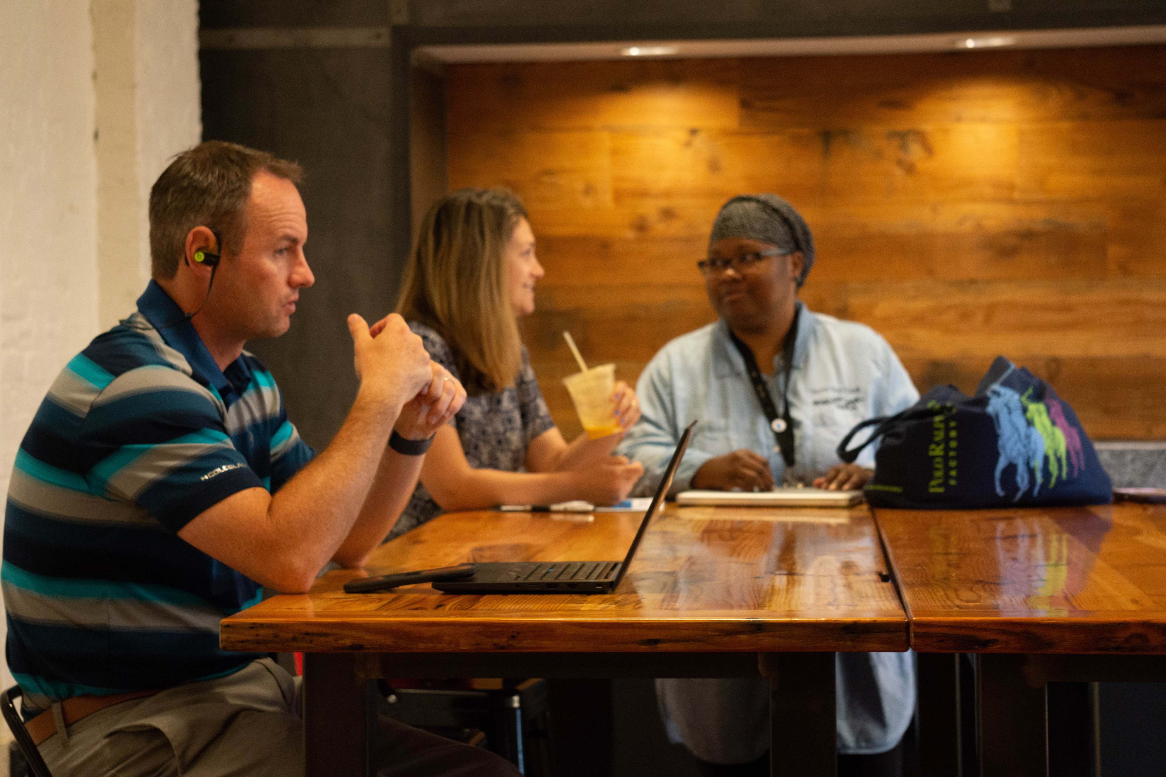 Three people are sitting at a wooden table in a cafe, with one person using a laptop and the others engaged in conversation.