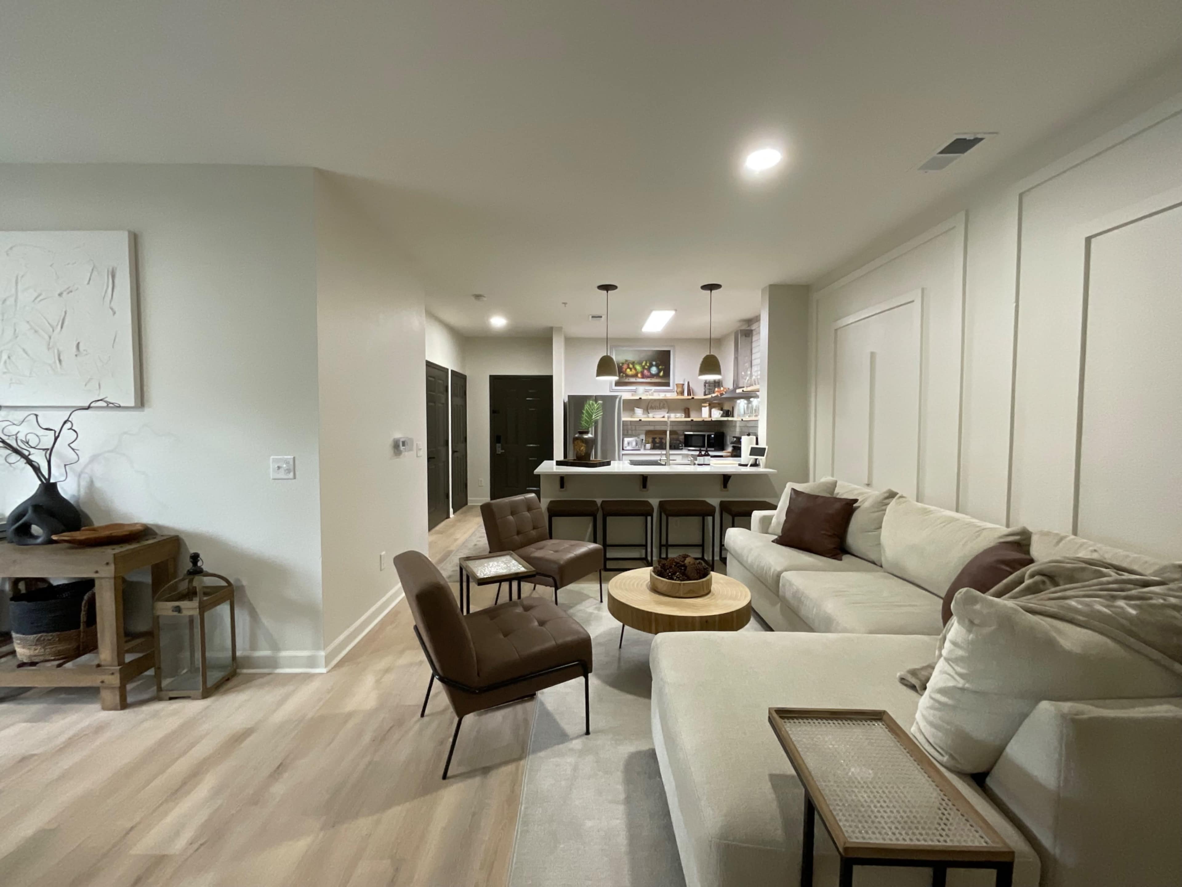 A modern living room features a light-colored sectional sofa, two brown chairs, and an open kitchen in the background with pendant lighting.