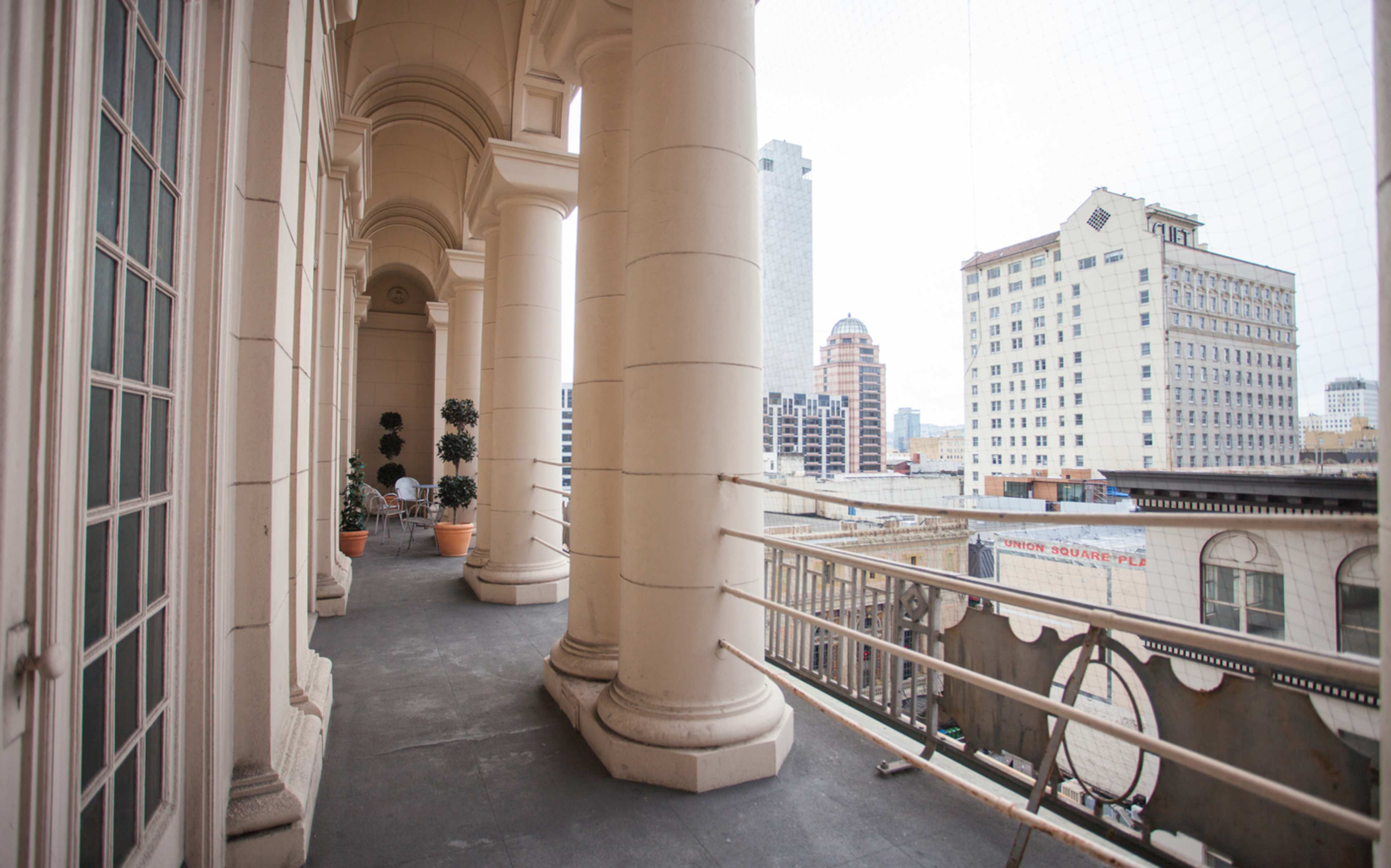 A balcony with large columns and potted plants, overlooking a cityscape with high-rise buildings in the background.