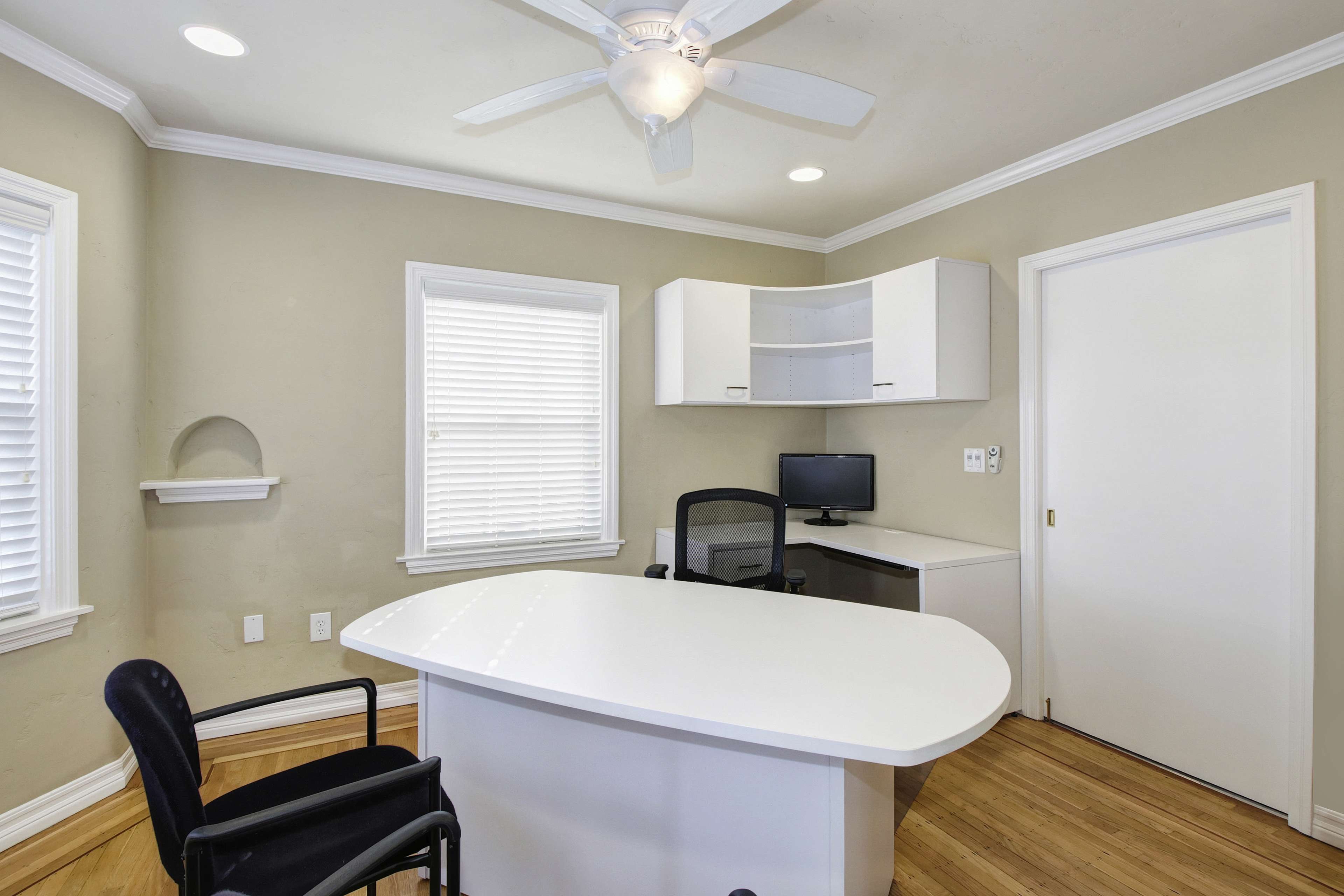 The image shows a neatly organized office space with a white desk, a black chair, a computer monitor, and natural light coming through the window.