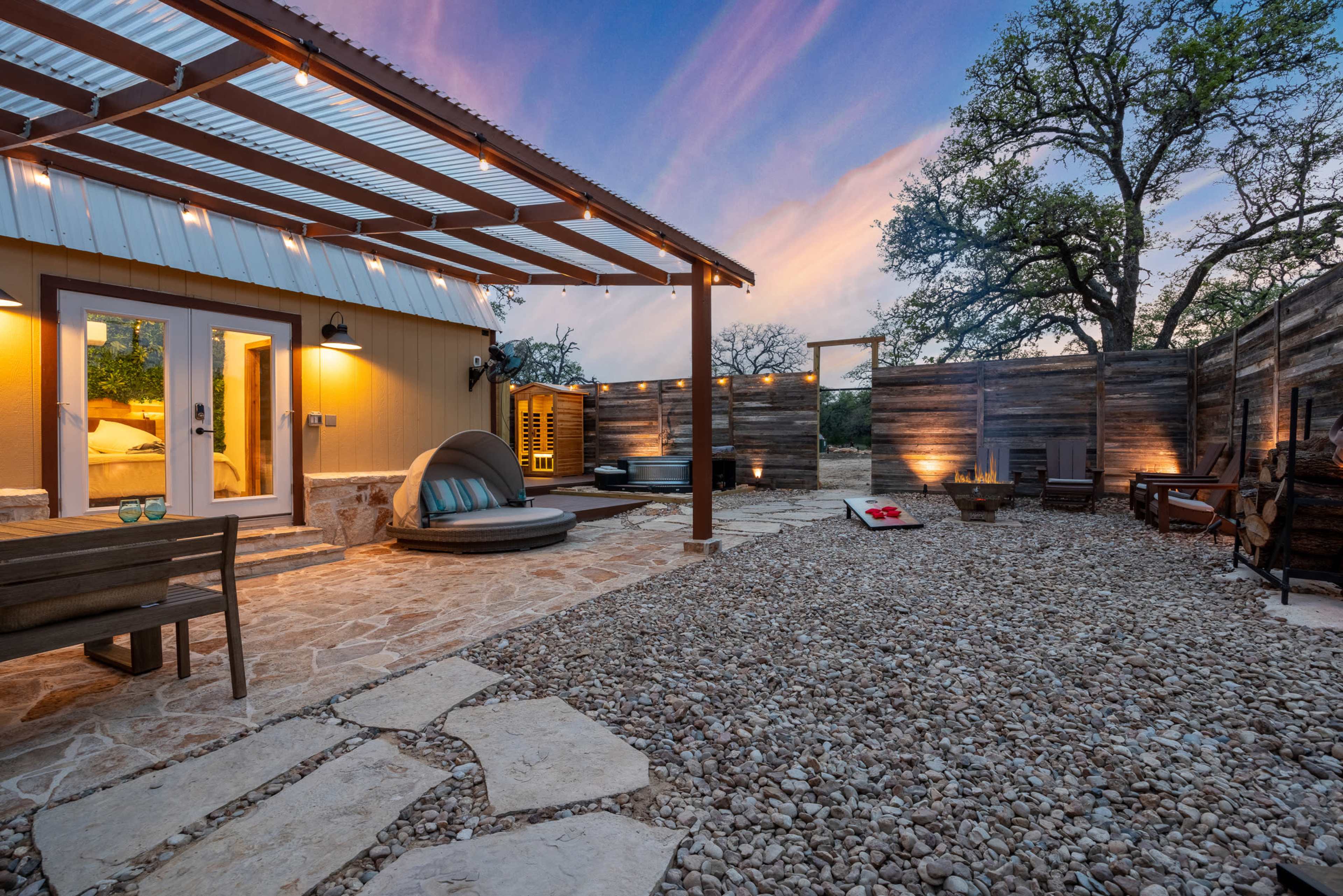 The outdoor area features a stone patio with a covered seating space, a curved lounge chair, and a wooden fence, all set against a colorful sunset sky.