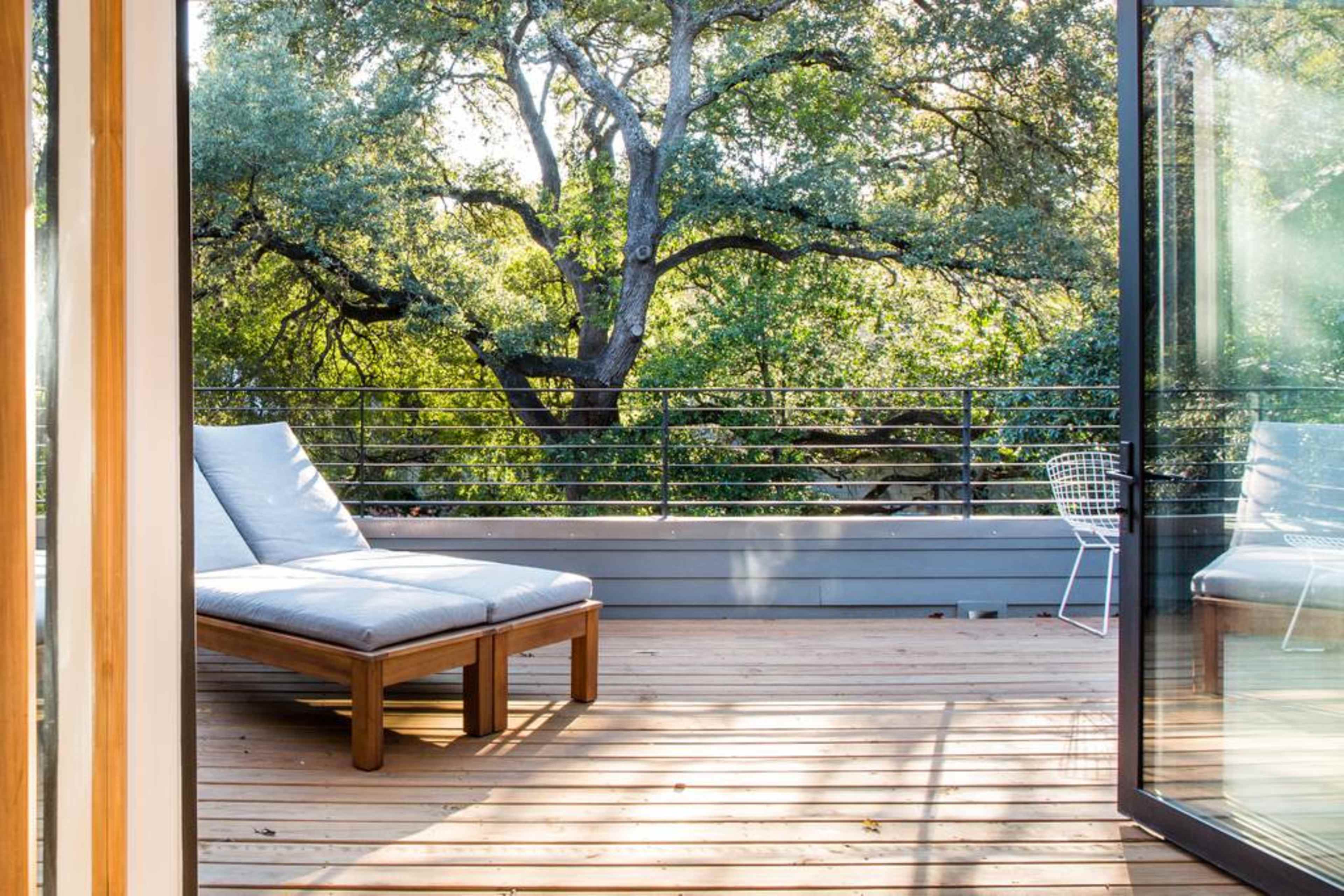 A wooden balcony features a lounge chair and a small chair, overlooking lush trees and greenery.
