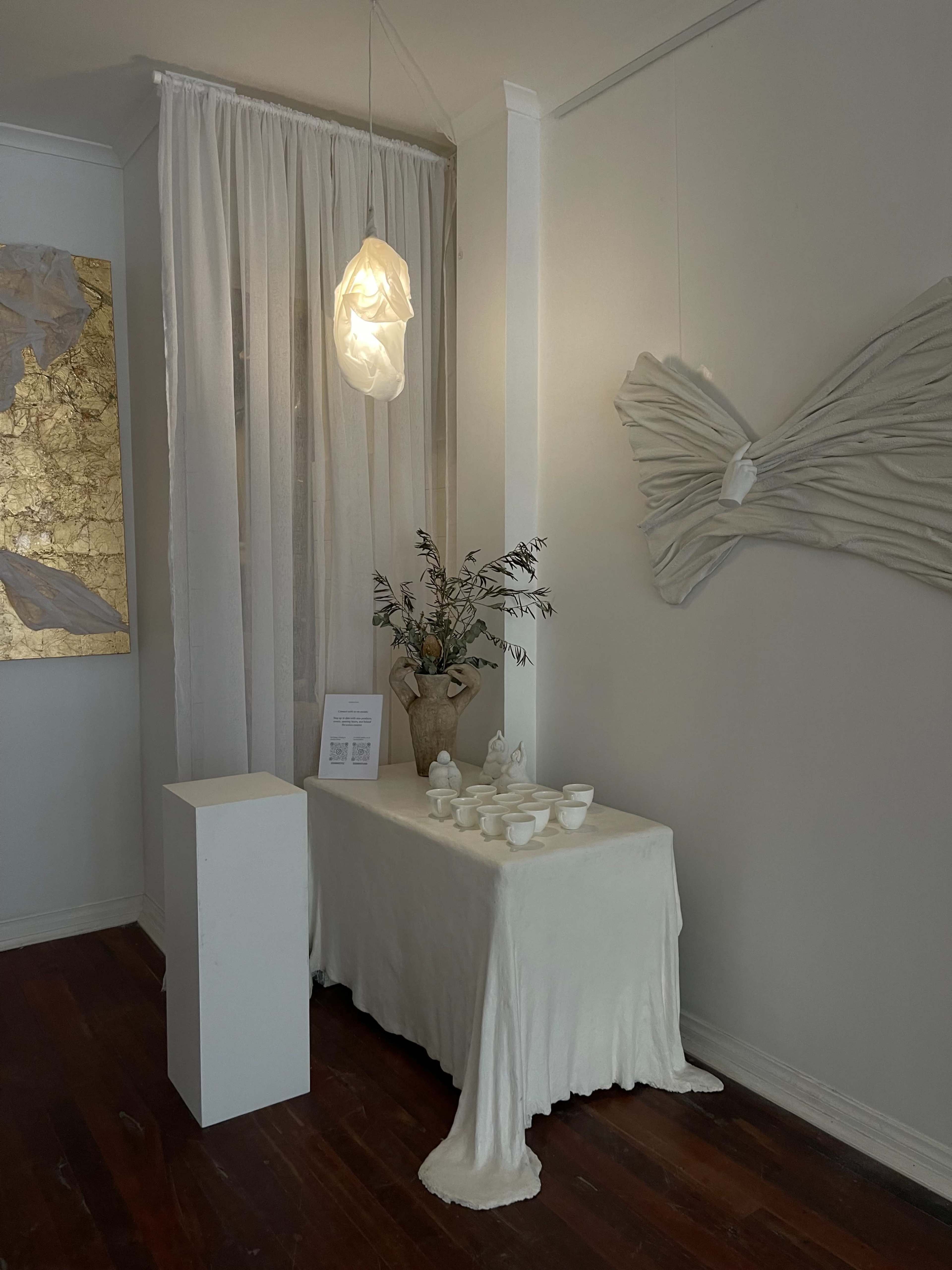The image shows a minimalist interior space with a table covered in a white tablecloth, adorned with ceramic cups, a vase of flowers, and a light fixture hanging above, adjacent to draped curtains and textured wall art.
