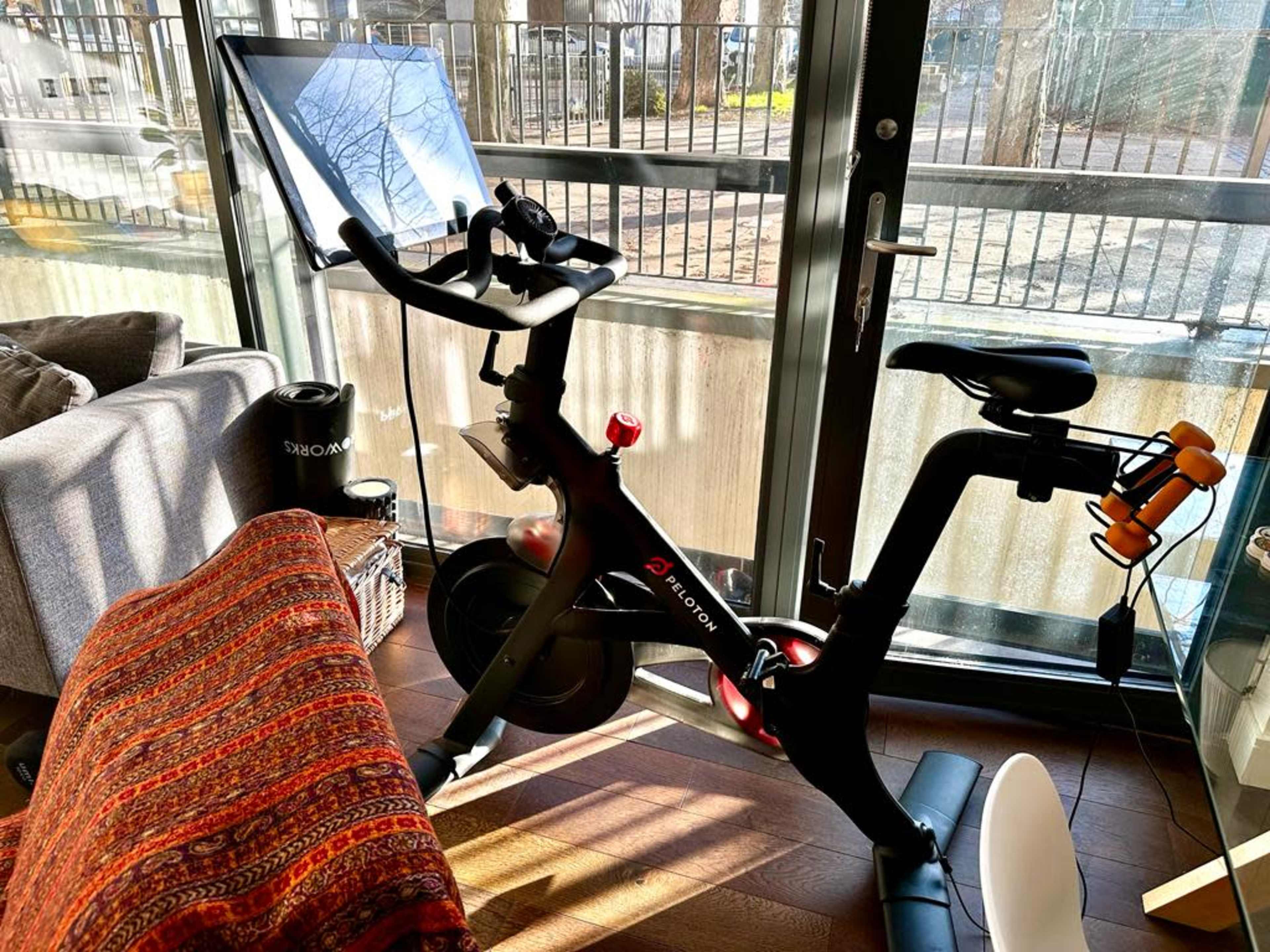 A stationary bike with a screen is positioned beside a window in a room with a couch and a colorful blanket.