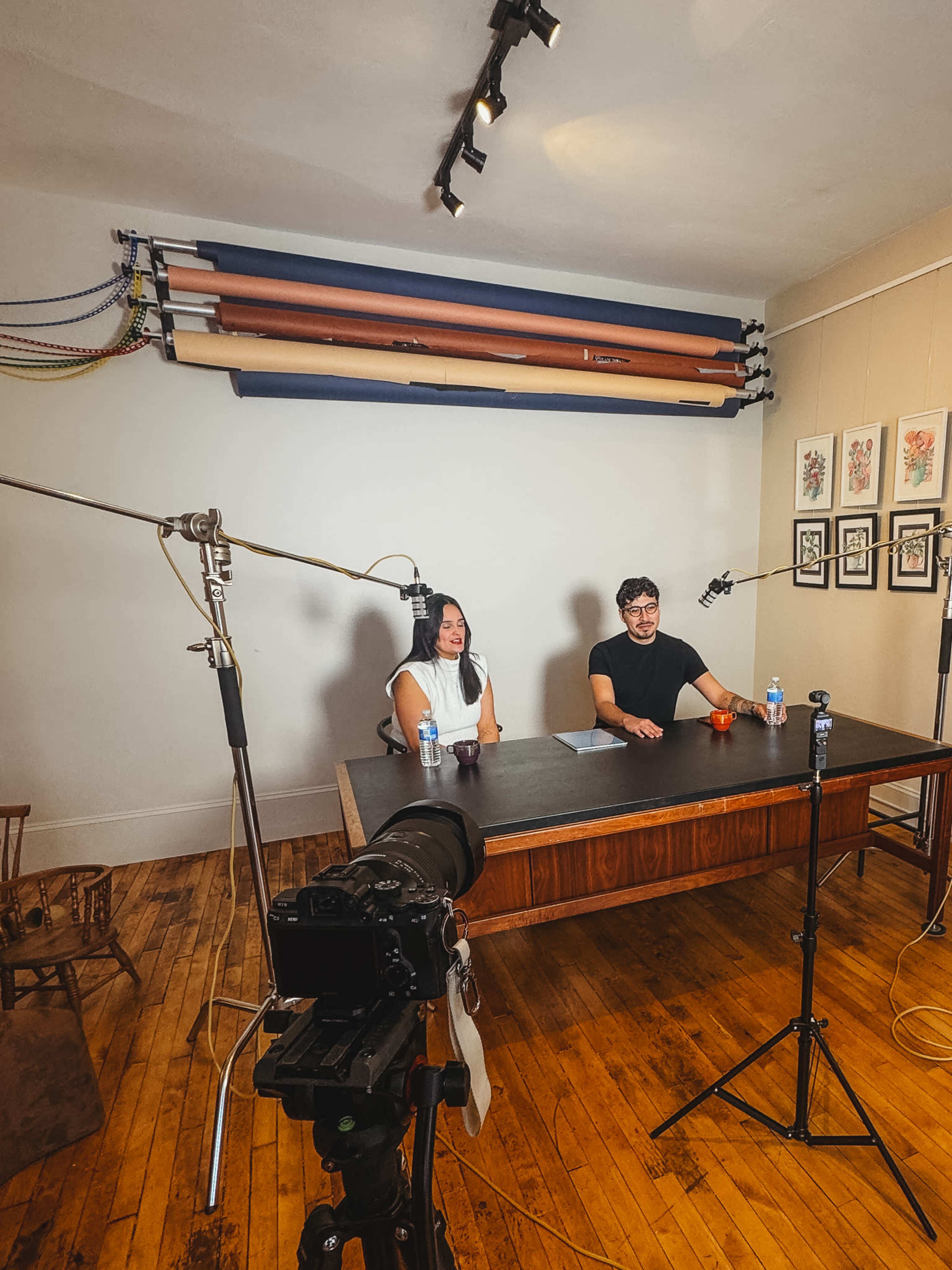 Two individuals sit at a large wooden table in a studio setting, with cameras and microphones set up for recording.