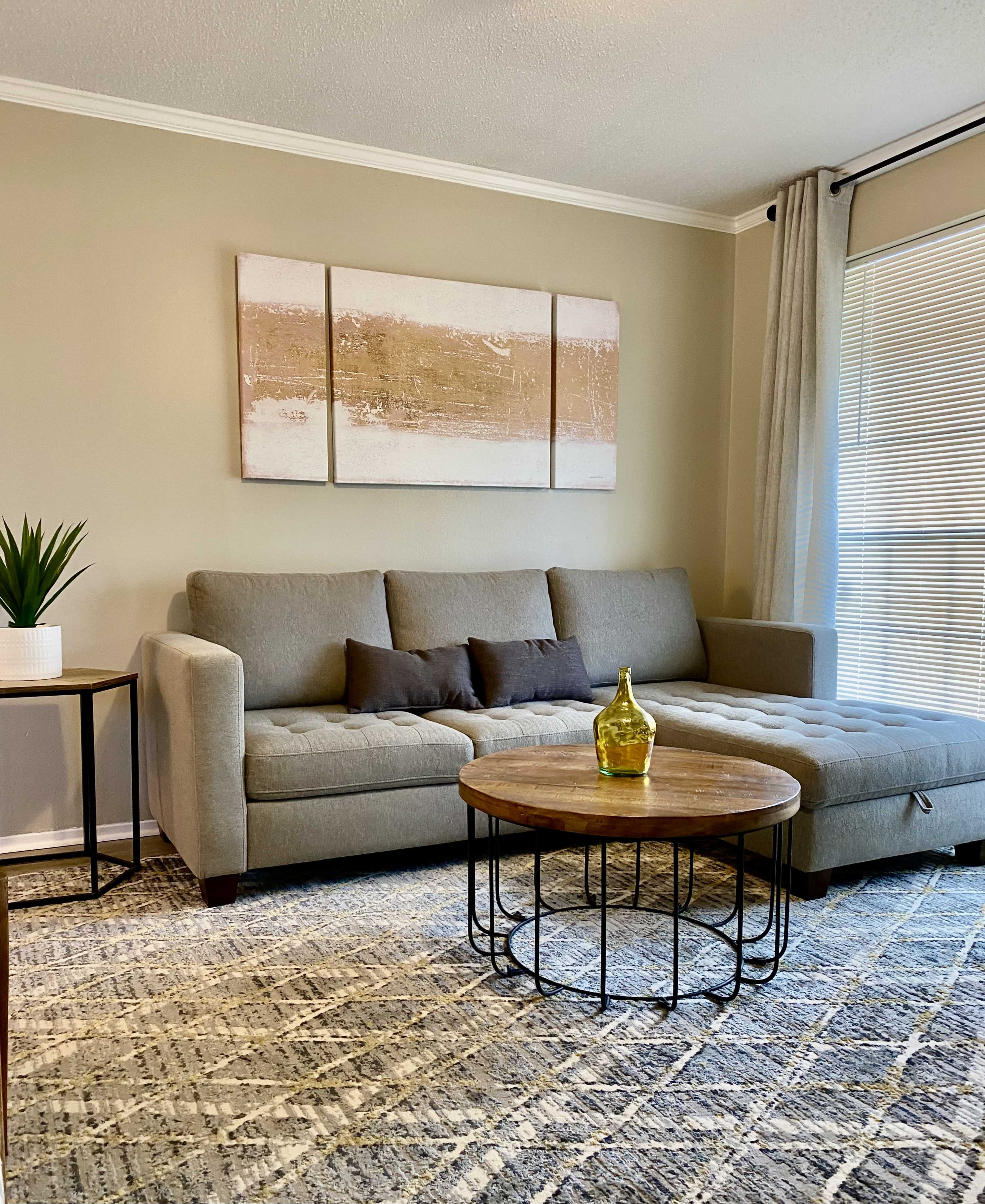 The scene features a modern living room with a gray sofa, a round wooden coffee table, and a decorative plant beside a window.