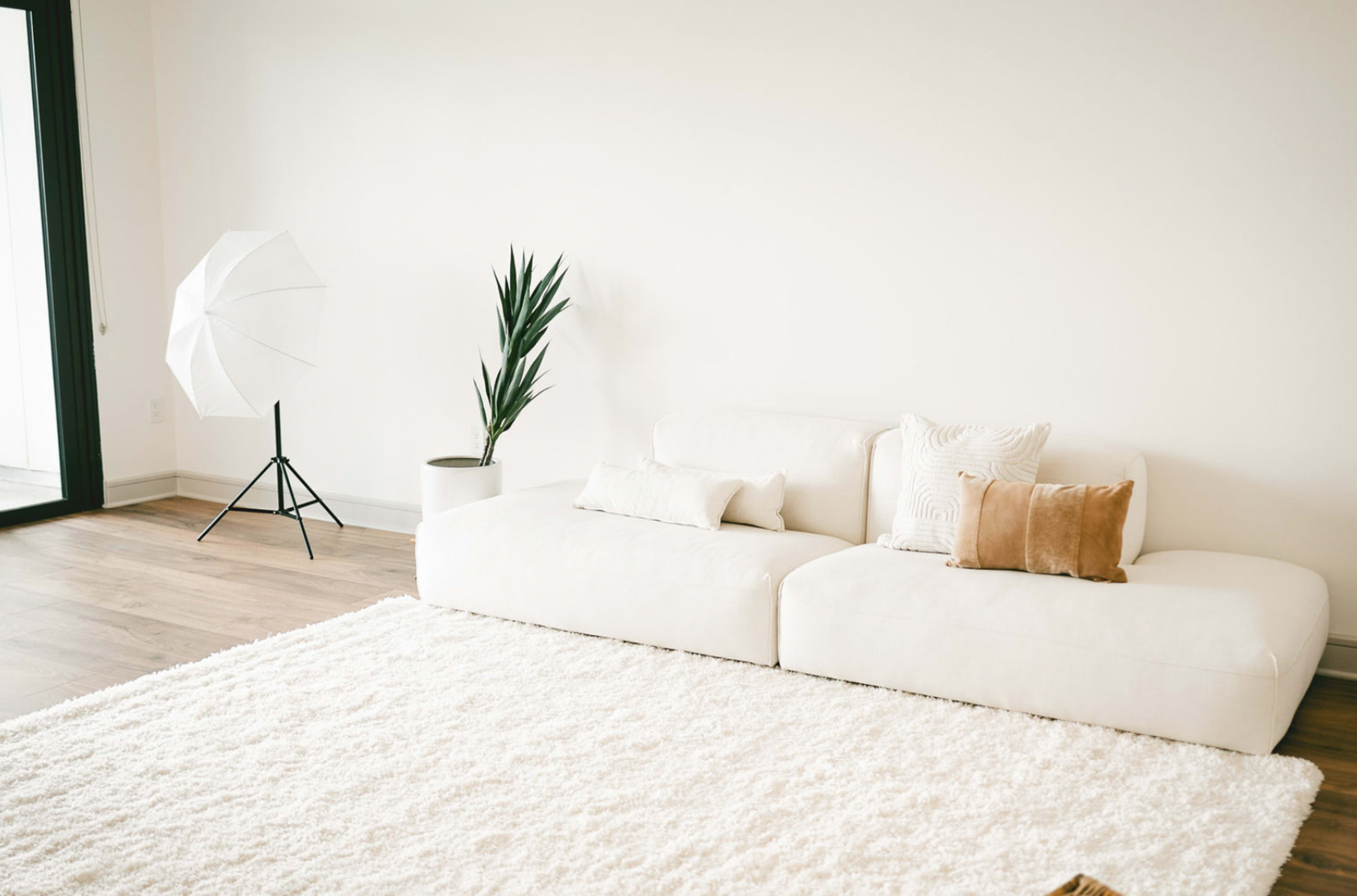 A white sectional couch sits on a fluffy rug in a minimalist room with a potted plant and a photography umbrella stand nearby.