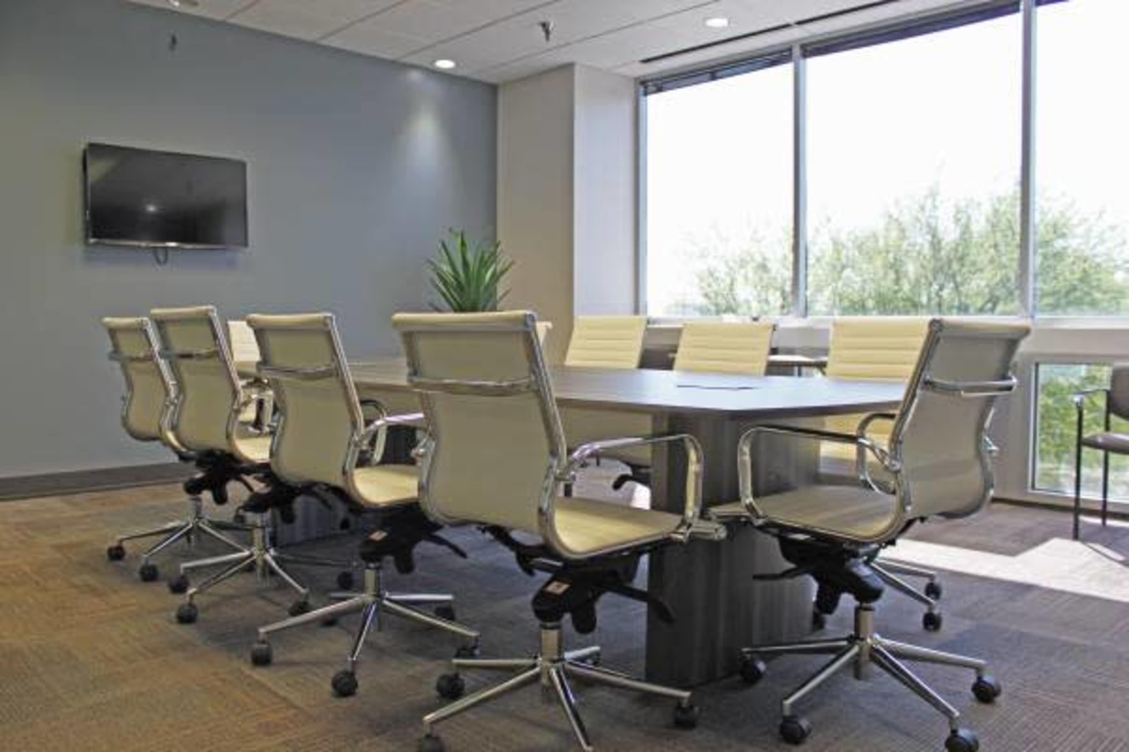A modern conference room features a large table surrounded by ergonomic chairs, with a television mounted on the wall and windows offering daylight.