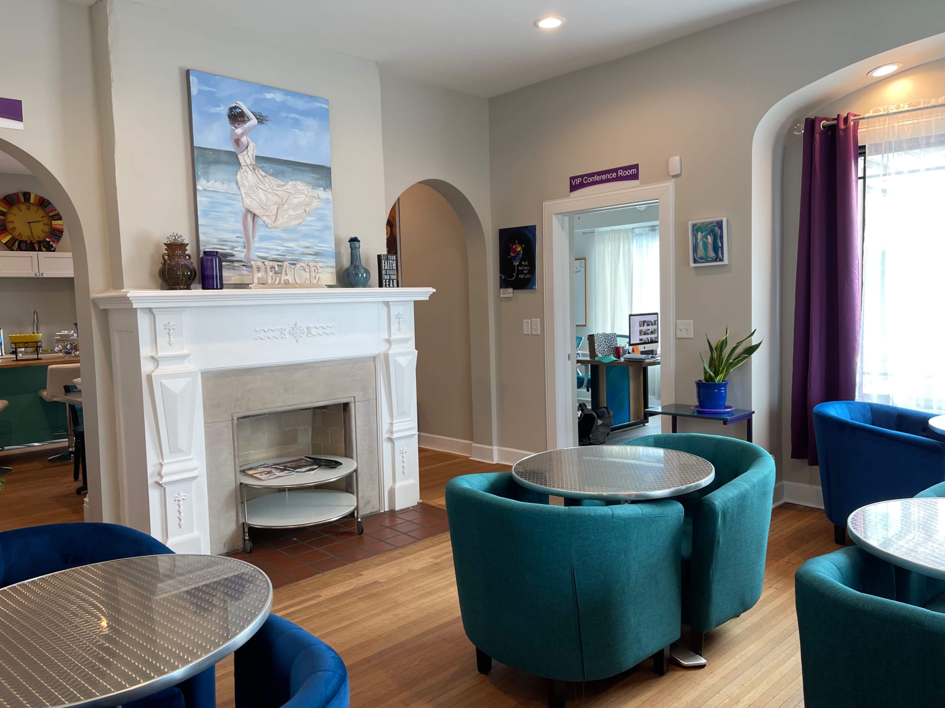 The image shows a cozy room with teal-colored chairs, a fireplace with a decorative mantle, and a painting depicting a woman by the sea.