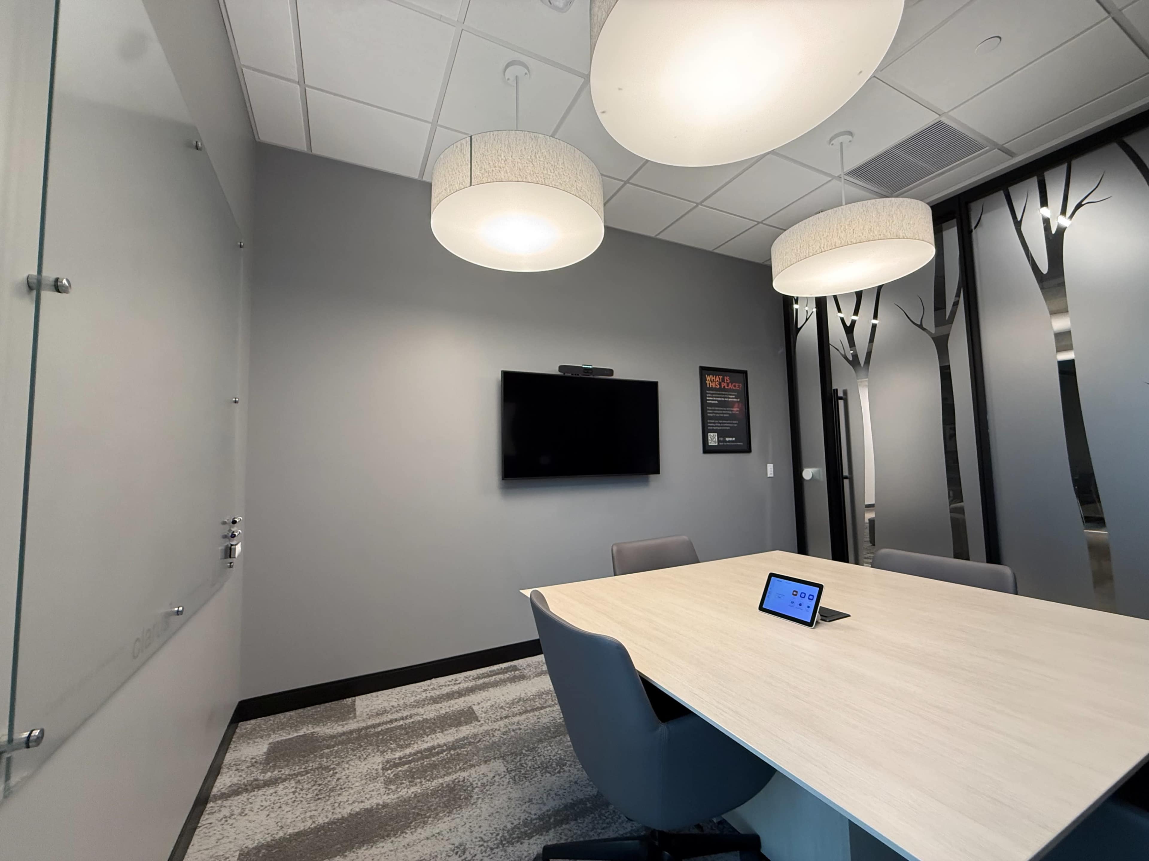The image shows a modern conference room with a large table, a wall-mounted television, an iPad on the table, and overhead lighting.