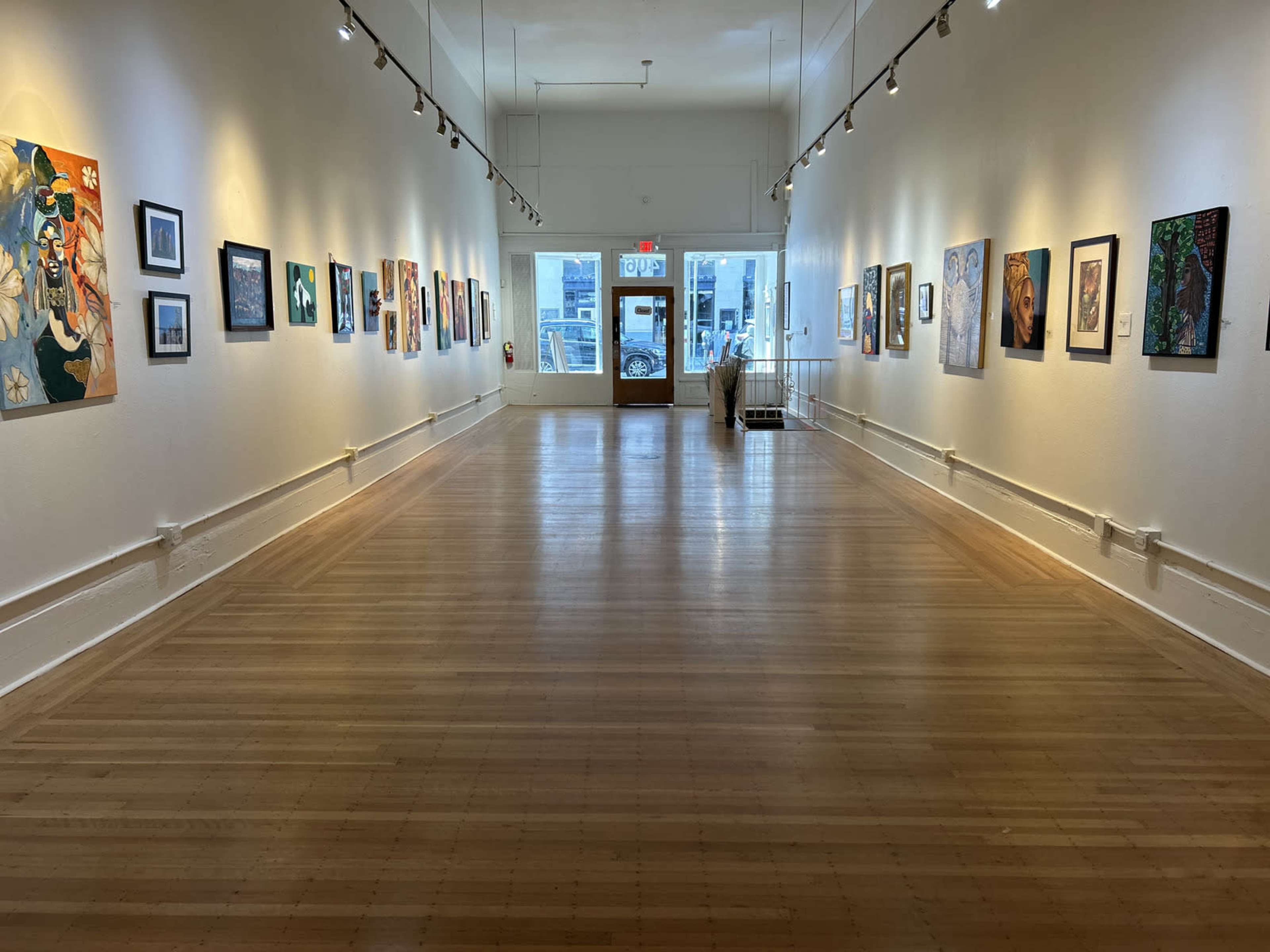 The image shows a well-lit art gallery featuring a long, empty hall lined with colorful paintings on the walls, leading to a bright entrance at the far end.