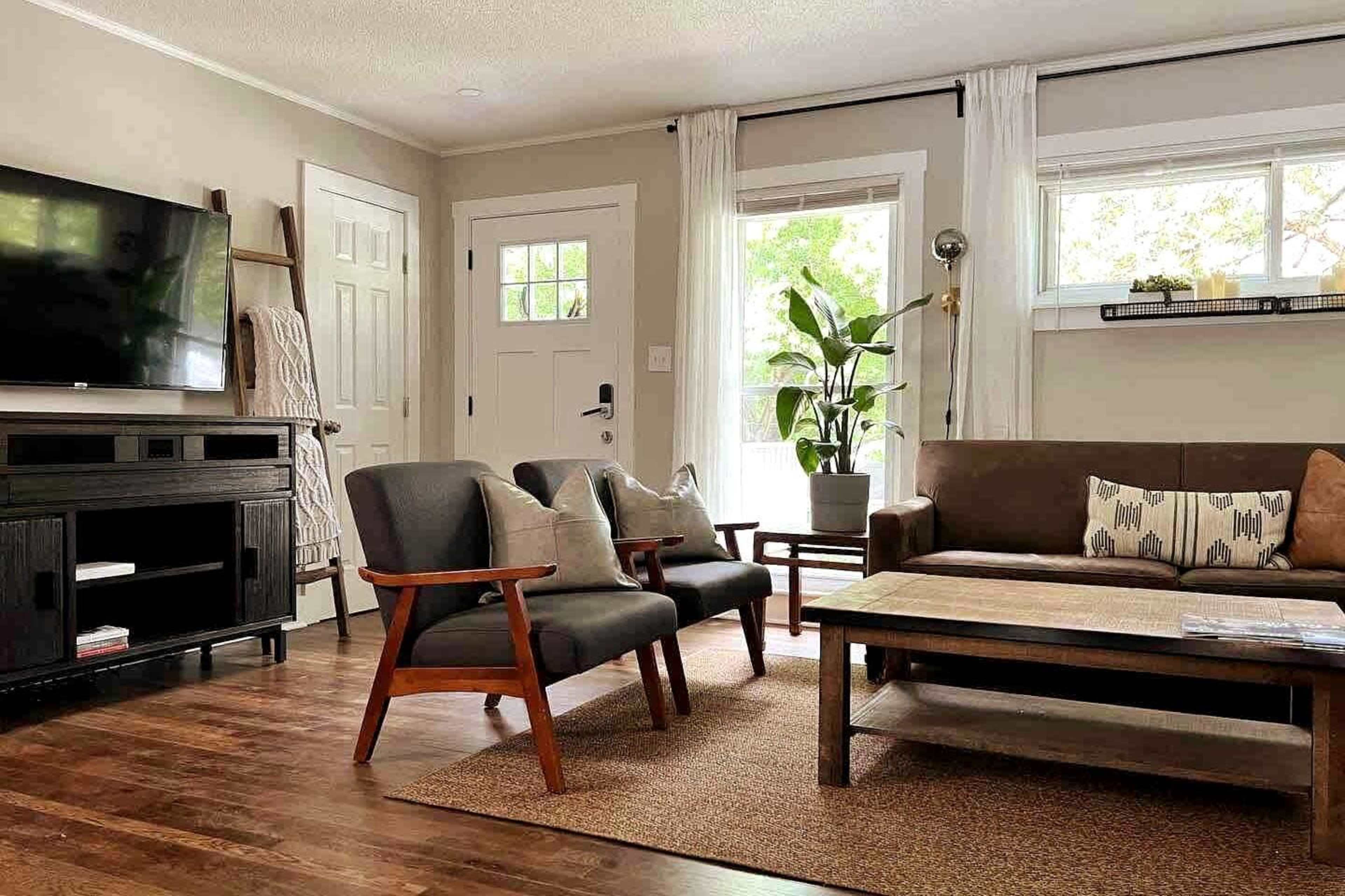 A cozy living room with a black television, two gray chairs, a brown couch, and a coffee table, all set on hardwood flooring with a large window letting in natural light.