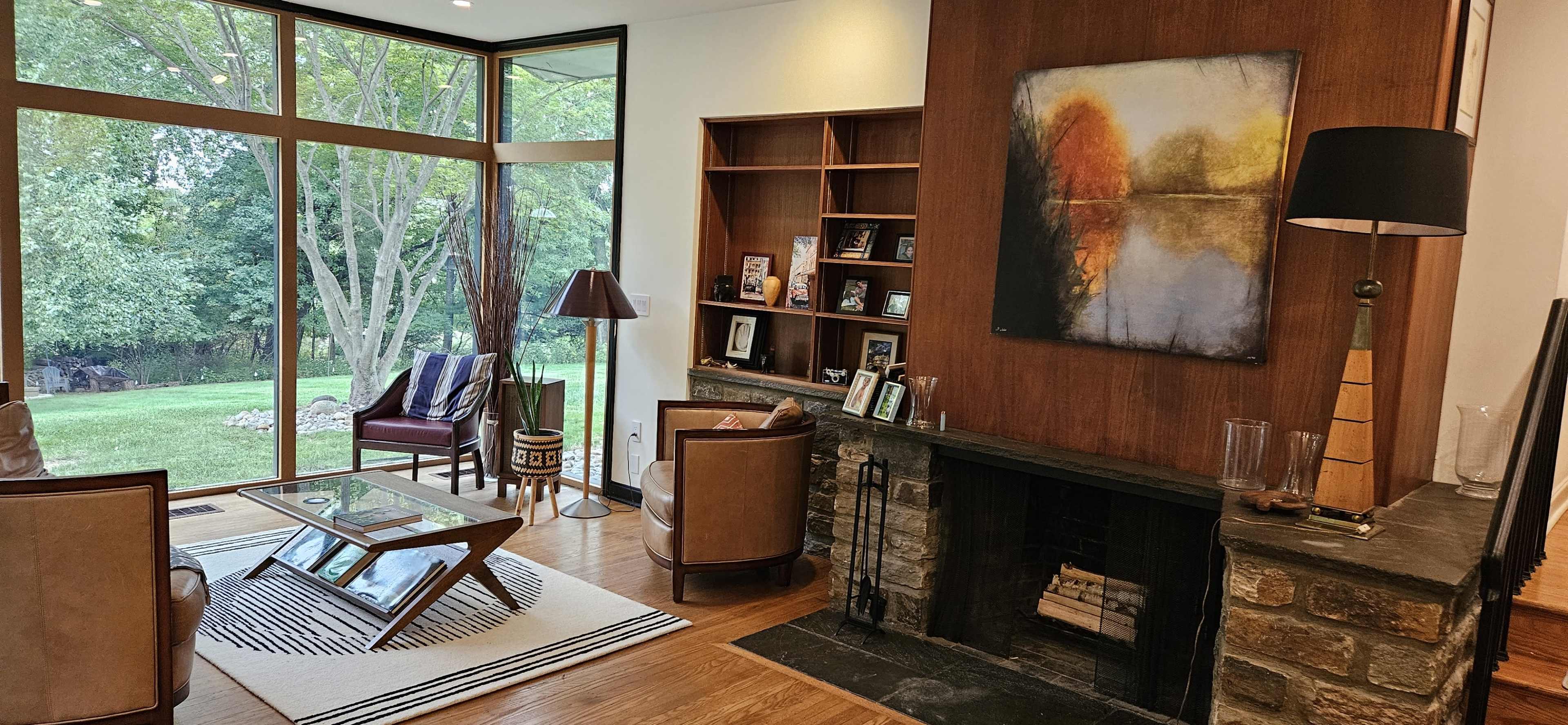 The image shows a modern living room with two armchairs, a coffee table, and a fireplace, all positioned near a large window that offers a view of a green backyard.