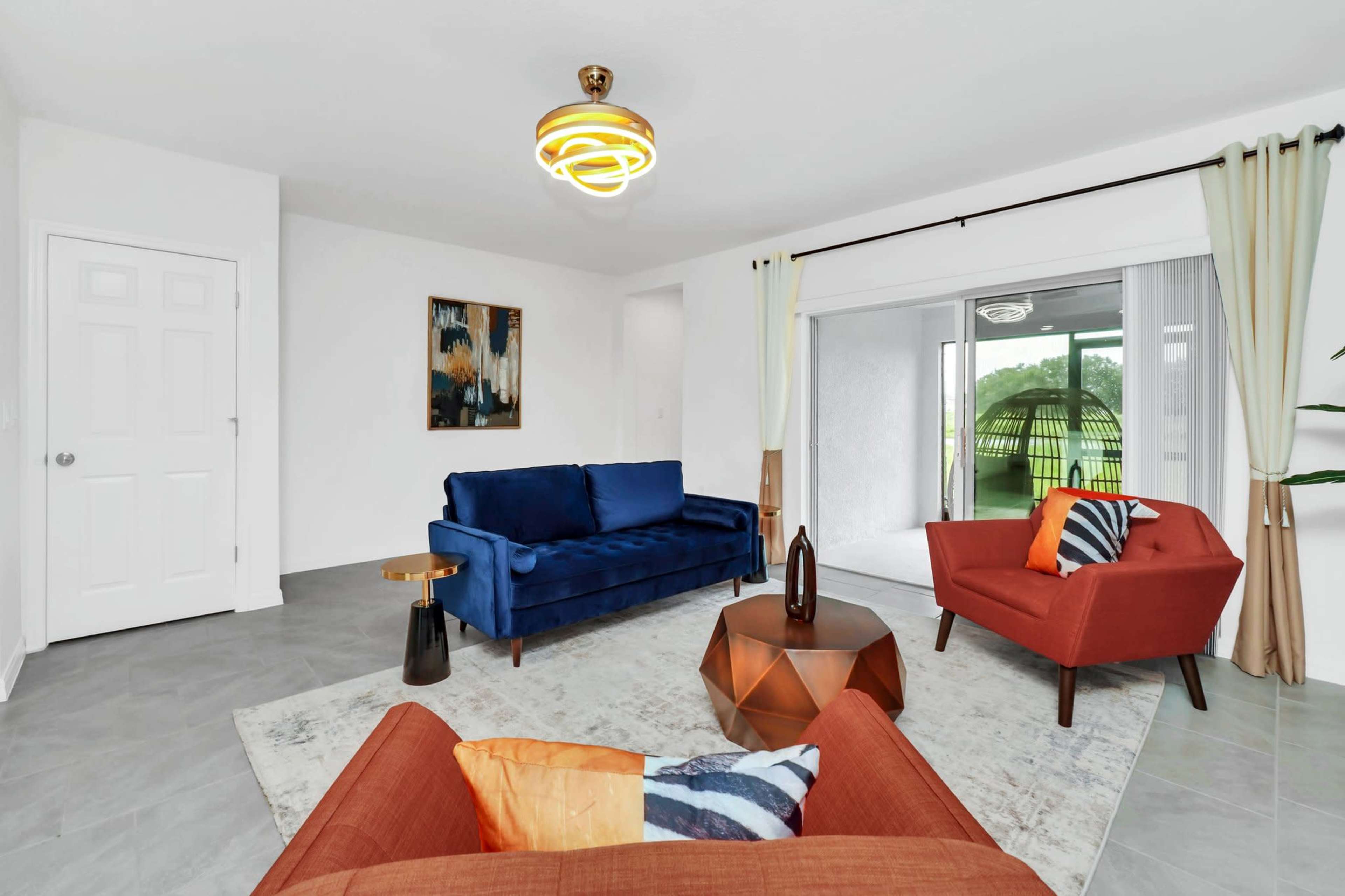 The room features a blue sofa and an orange armchair arranged around a geometric coffee table, with a sliding door providing access to an outdoor area.