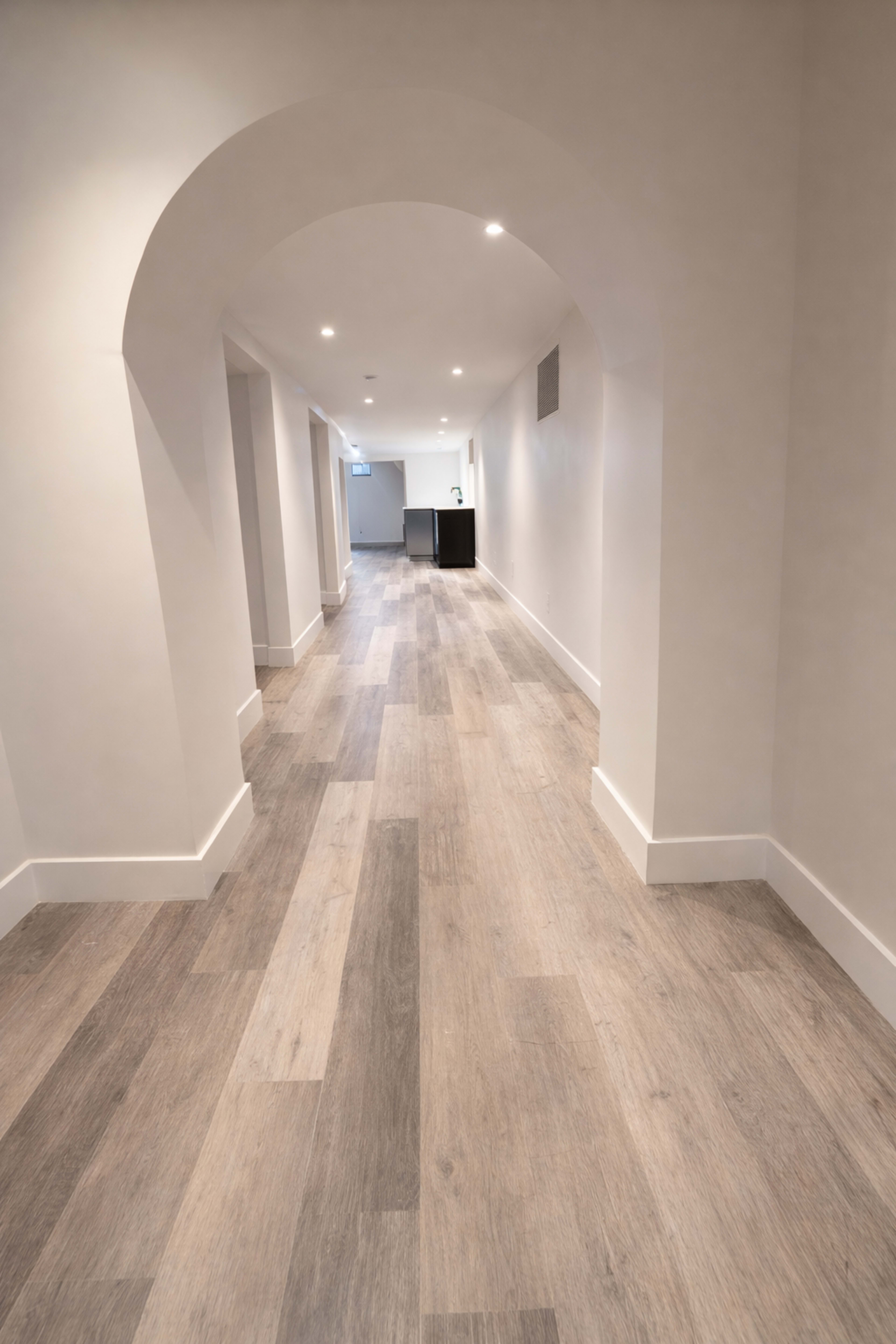 The image shows a long, narrow hallway with a curved archway at one end and light-colored wooden flooring.