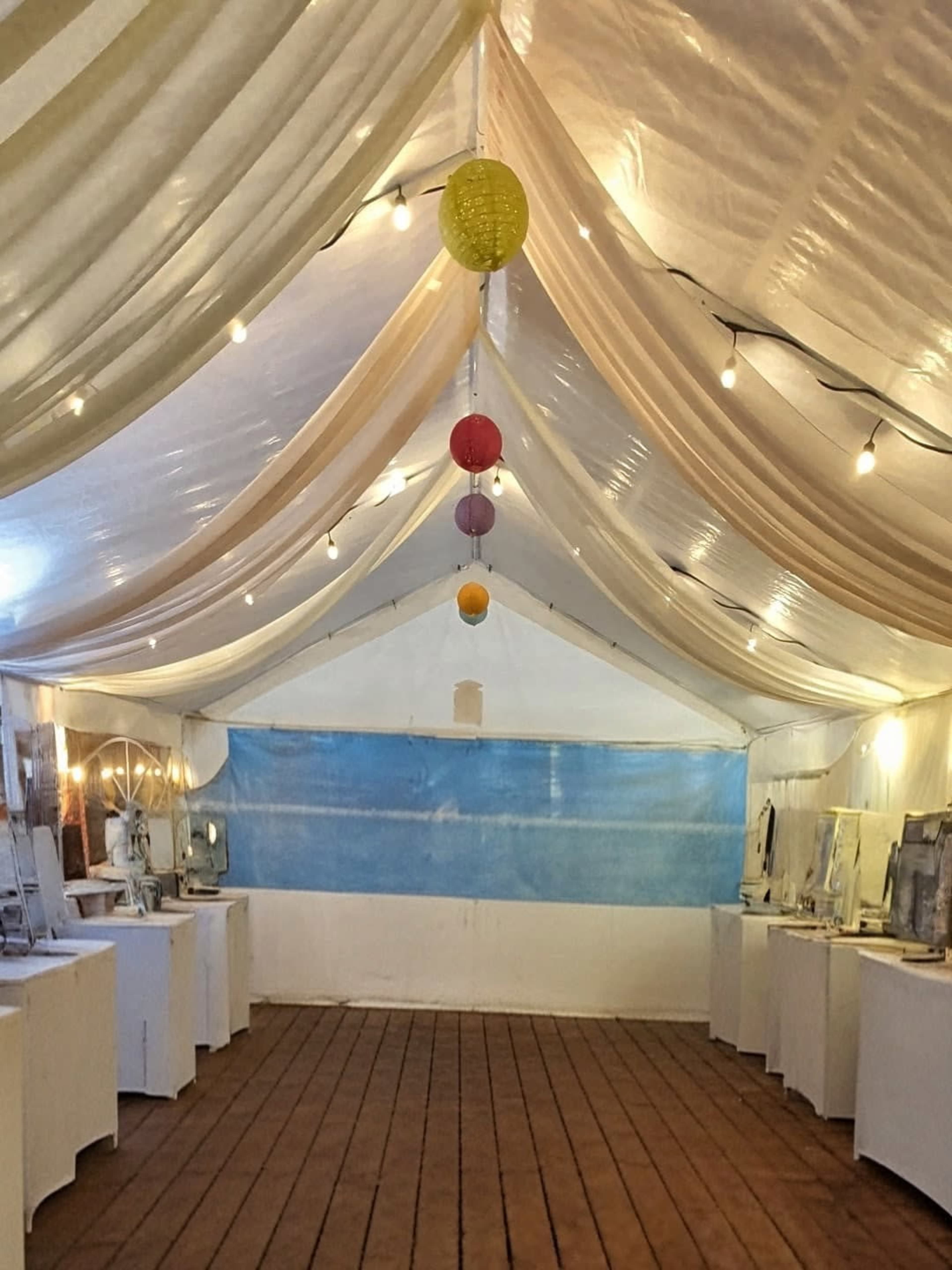 The image shows a tented space with draped fabric overhead, wooden flooring, and several tables lined along the sides, illuminated by string lights and colorful hanging lanterns.