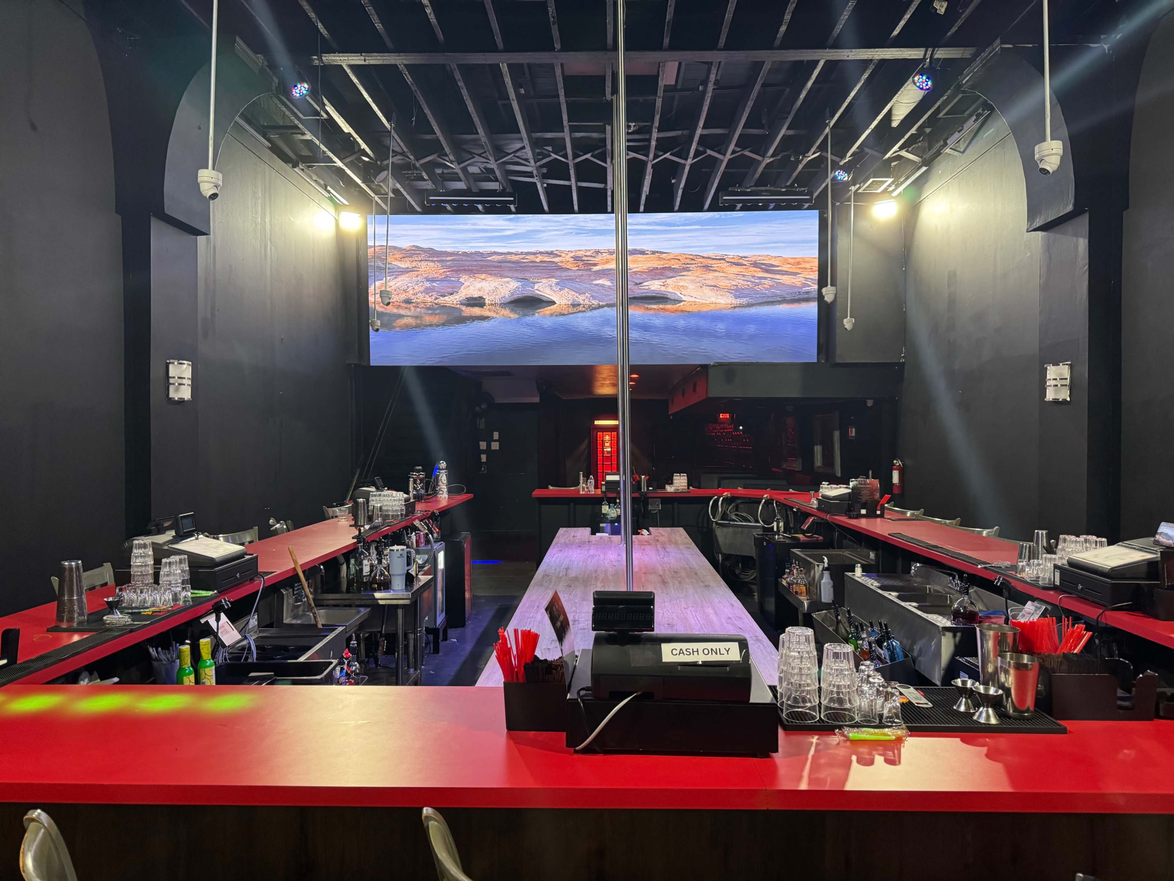 The image shows a bar area with red countertops, multiple bottles and glassware arranged on shelves, and a large screen displaying a landscape in the background.