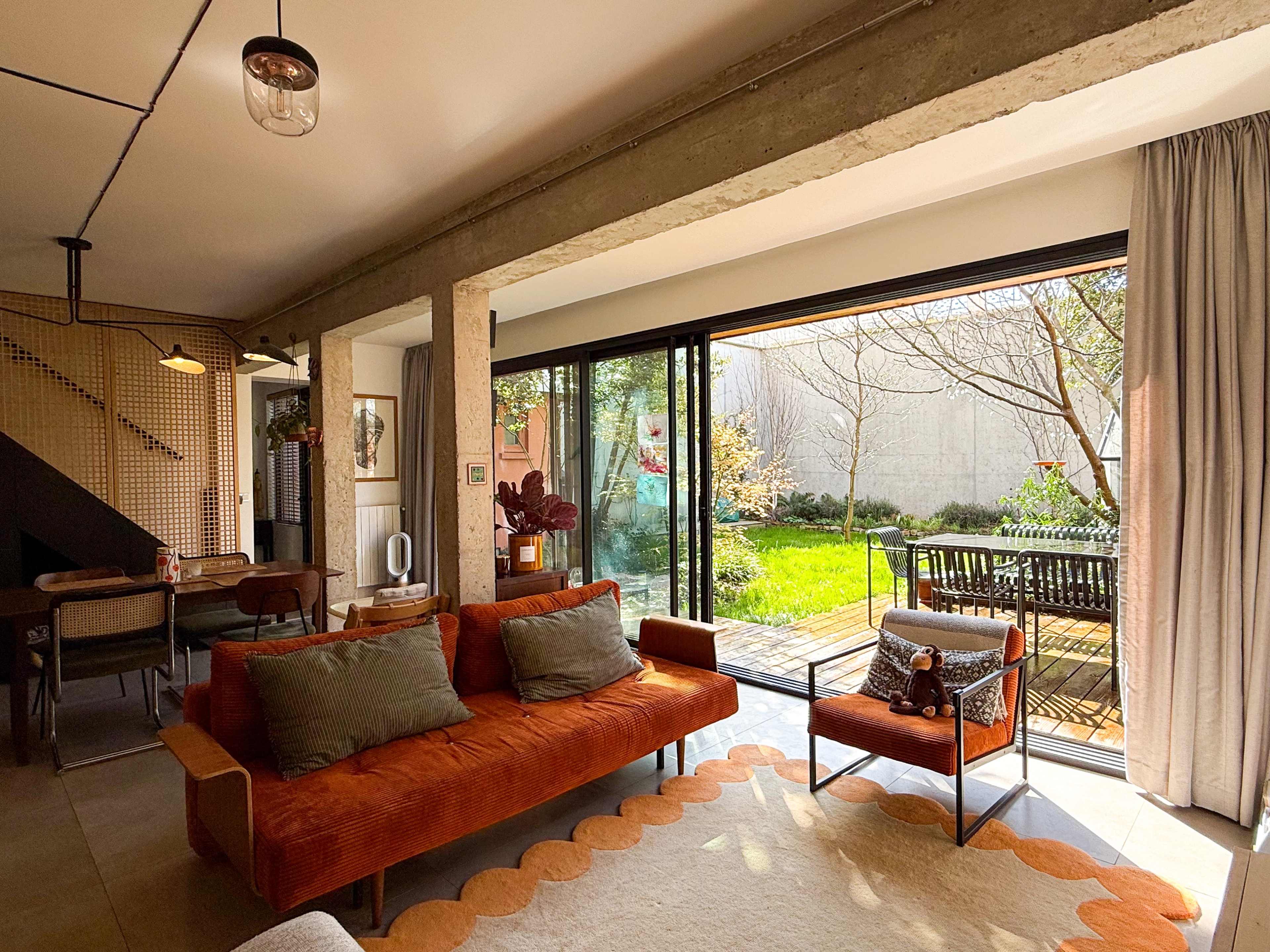 The image shows a modern living room with a brown couch and a small chair, facing large glass doors that open to a garden area.