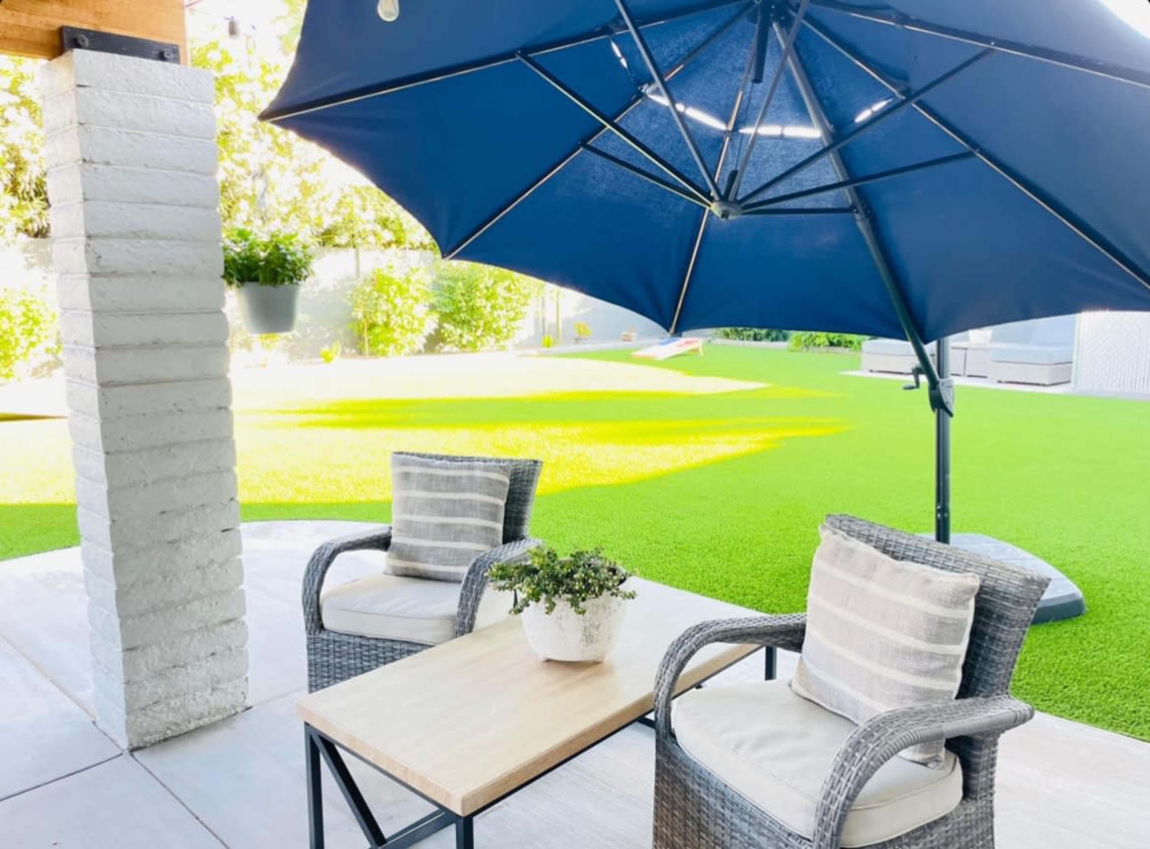 A patio area features two wicker chairs and a table beneath a large blue umbrella, with artificial grass visible in the background.
