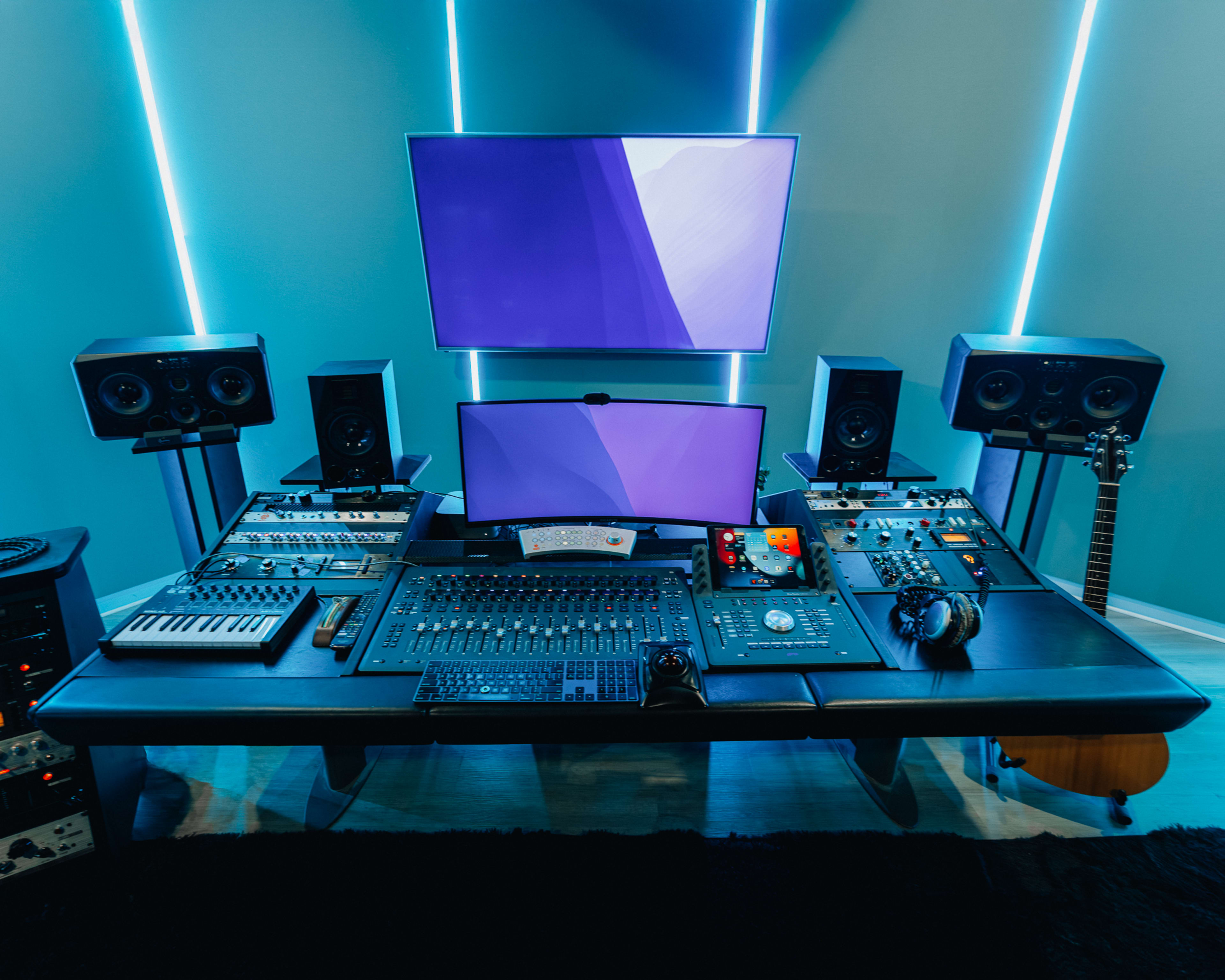 The image shows a modern music production studio with a large desk equipped with audio mixing equipment, a computer monitor, studio speakers, and a guitar.