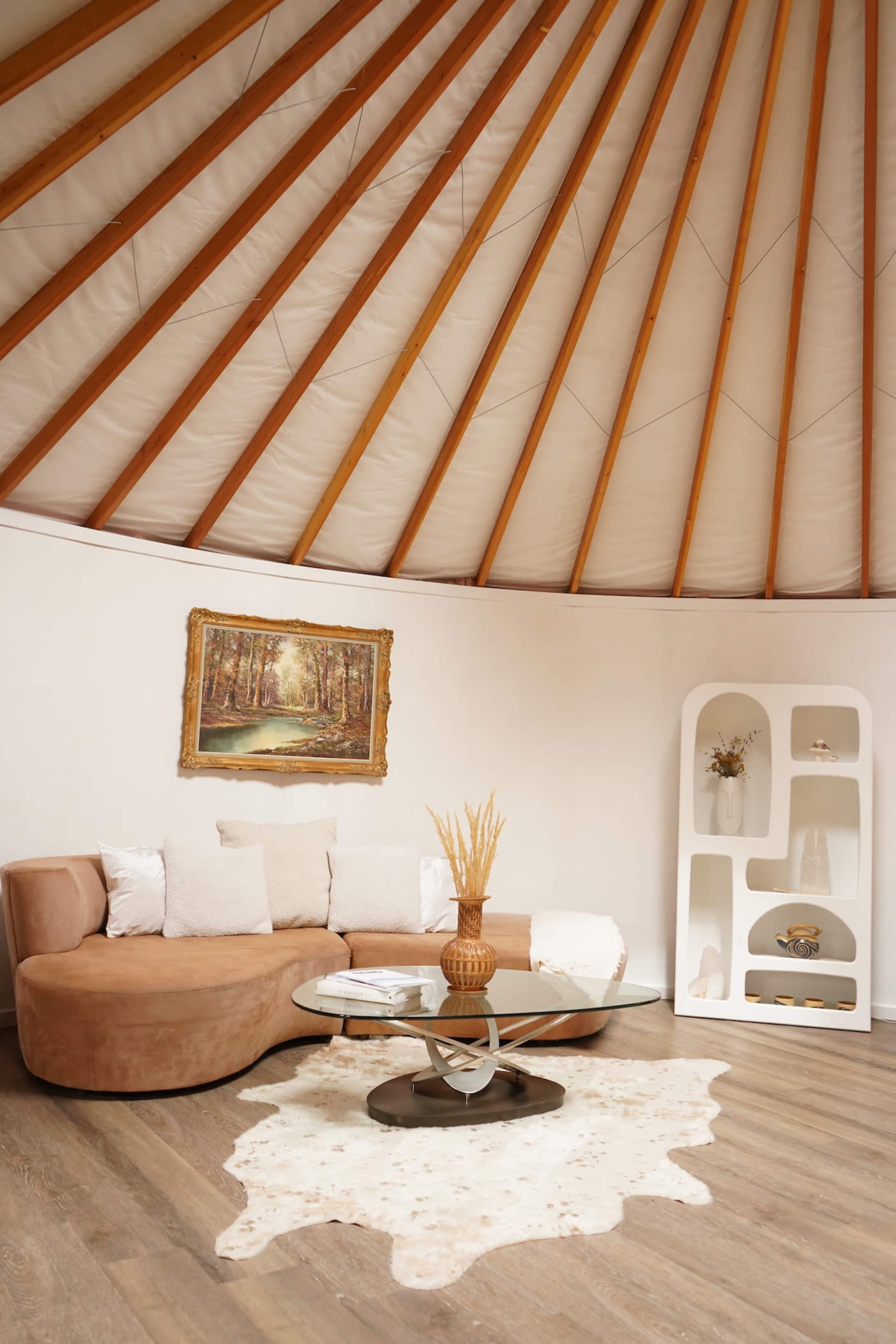 A modern circular room with wooden beams, featuring a curved brown sofa, a round coffee table, a decorative vase, and a framed painting of a forest.