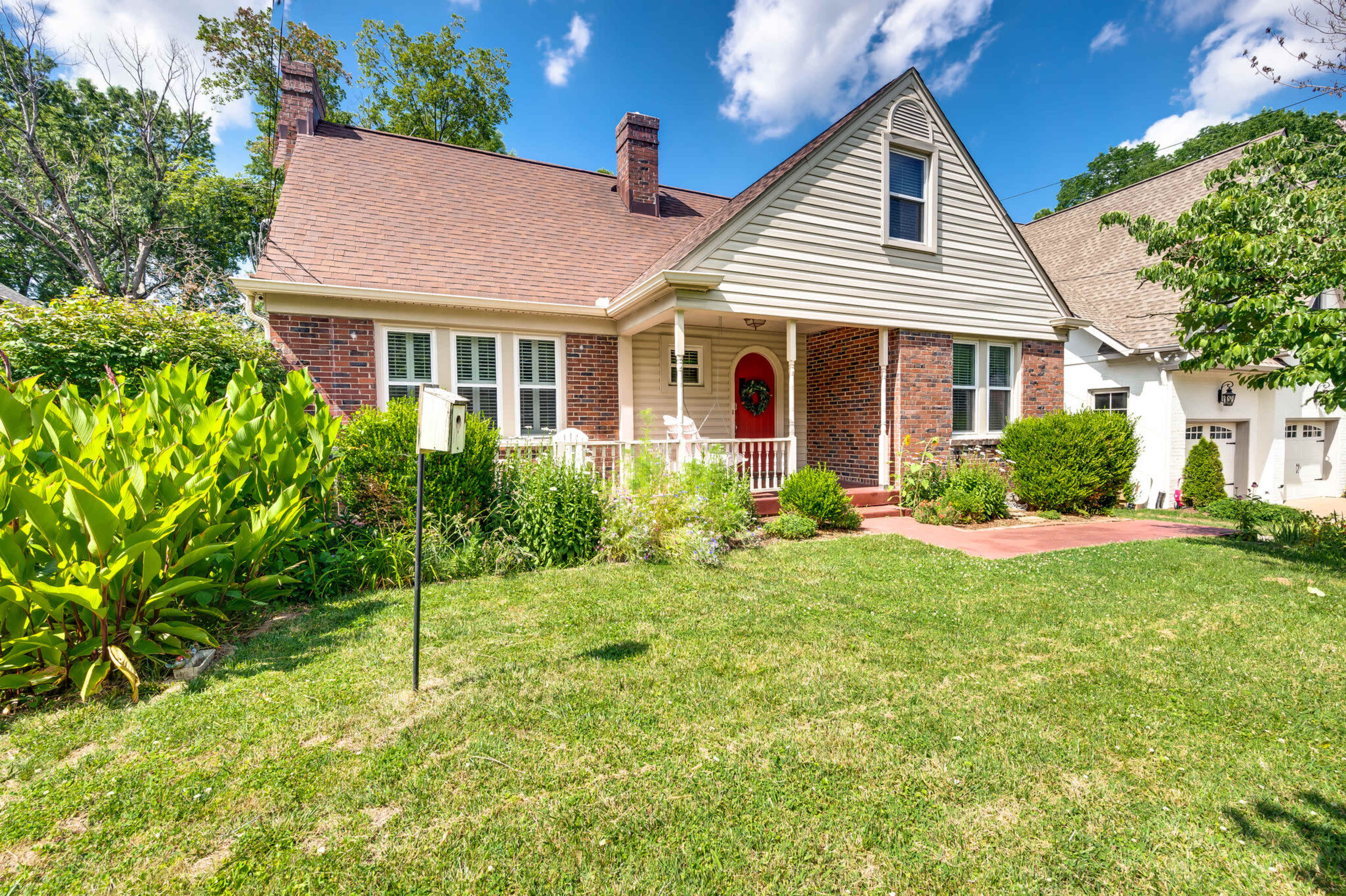 A brick house with a front porch and a red door is surrounded by green shrubs and grass under a blue sky.