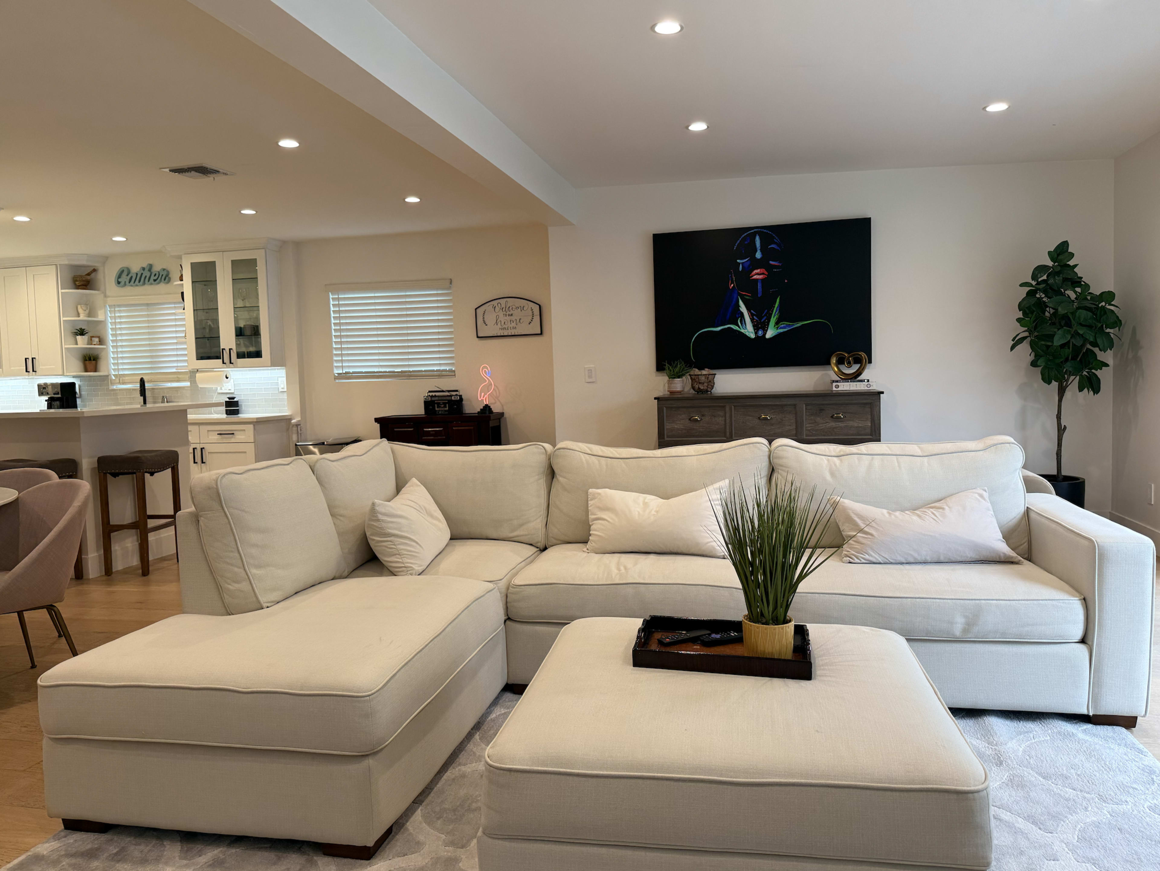 A light-colored sectional sofa with decorative pillows sits in a well-lit living room, featuring a modern artwork on the wall and a green potted plant nearby.