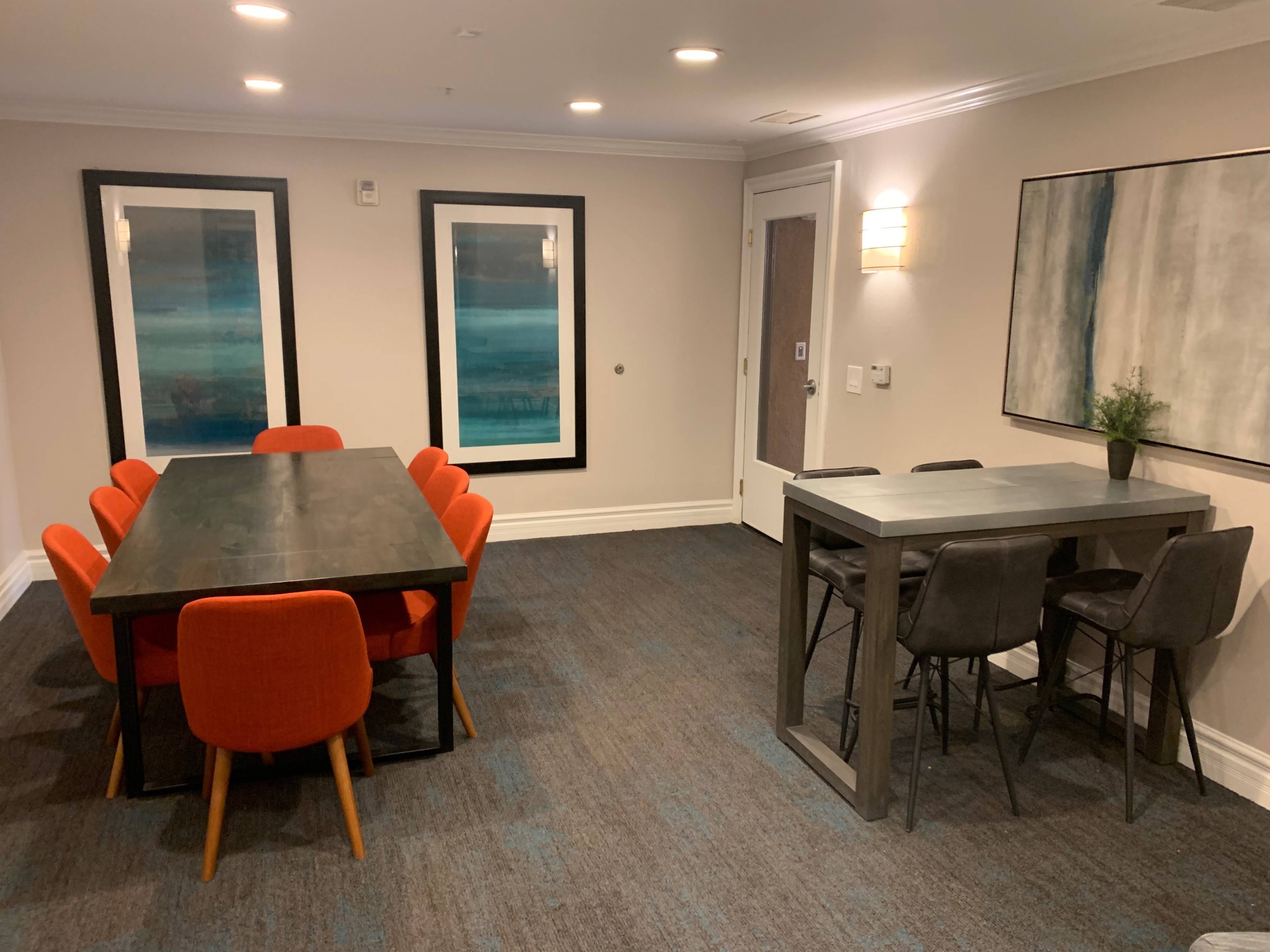 The image shows a modern room with a dining table surrounded by eight orange chairs, a small tall table with four dark chairs, and framed artwork on the walls.