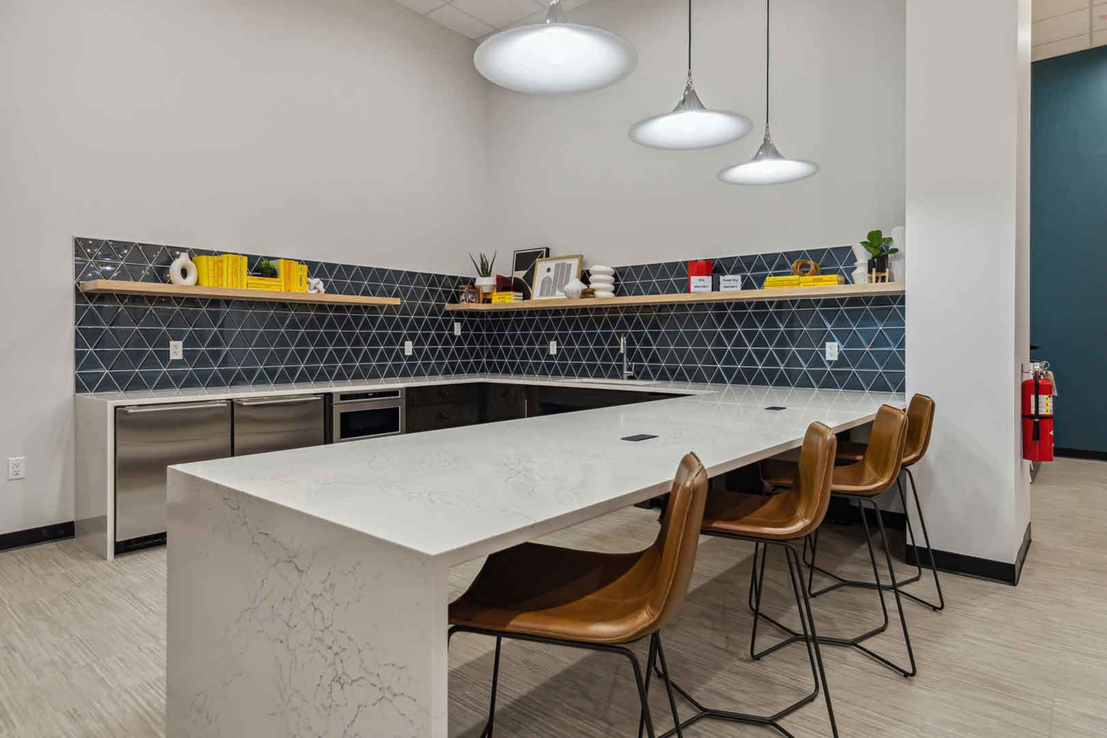 A modern kitchen with a large white countertop, four brown chairs, and geometric tile backsplash featuring decorative items on shelves.