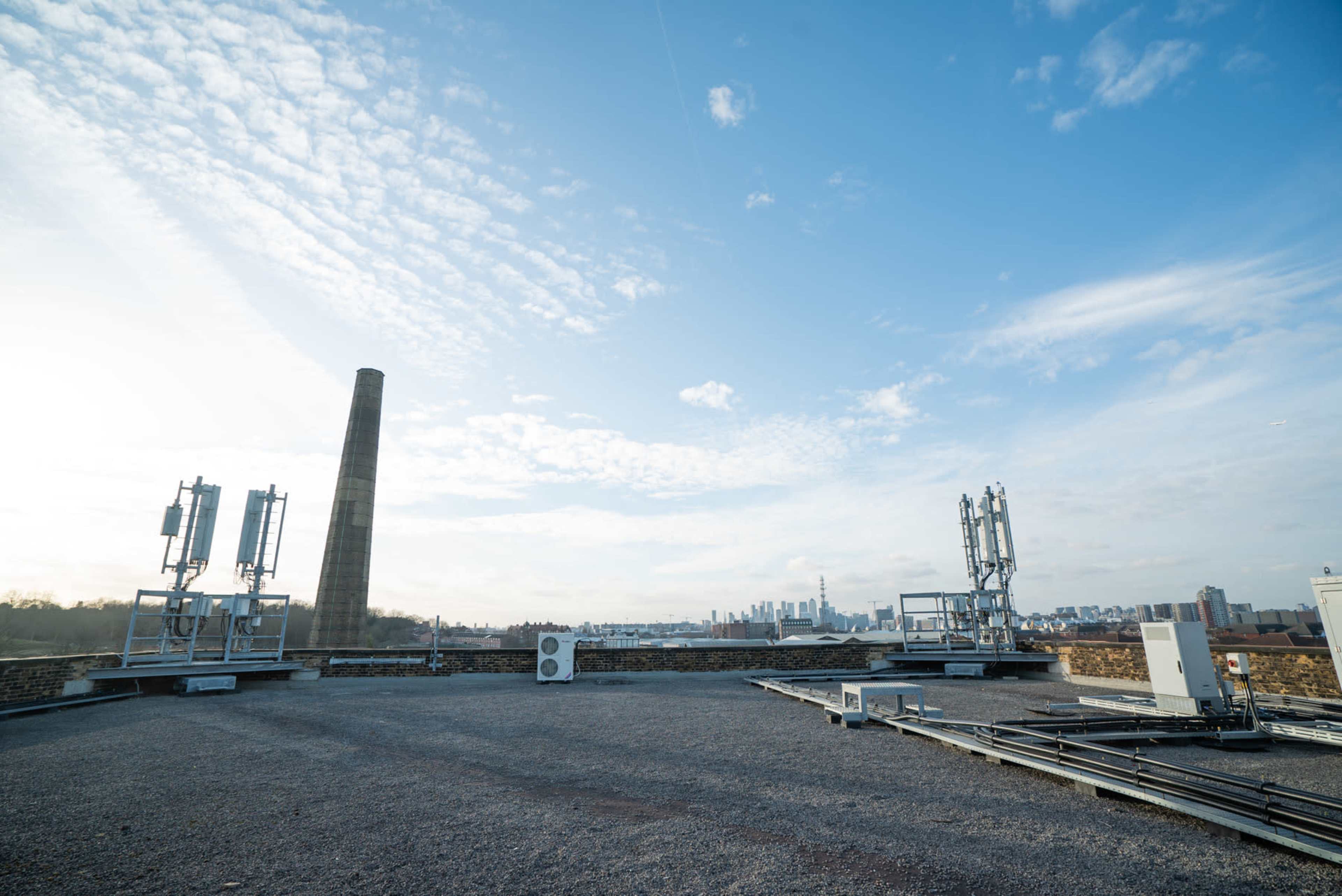 A rooftop with telecommunications equipment and a tall brick chimney, set against a cloudy sky and a distant skyline.