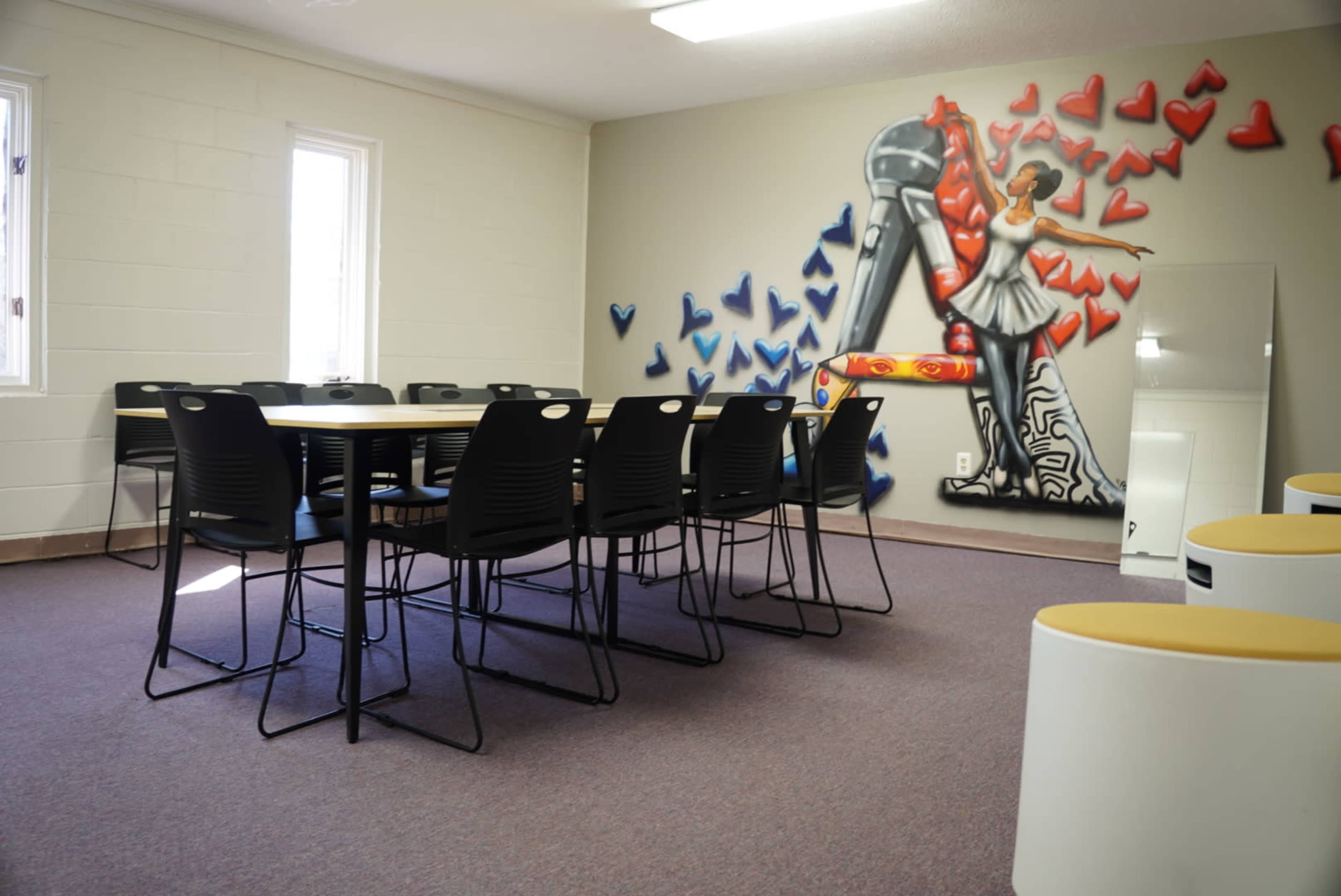 A room with a table surrounded by black chairs and a mural of a figure in front of a large red and blue heart design on the wall.