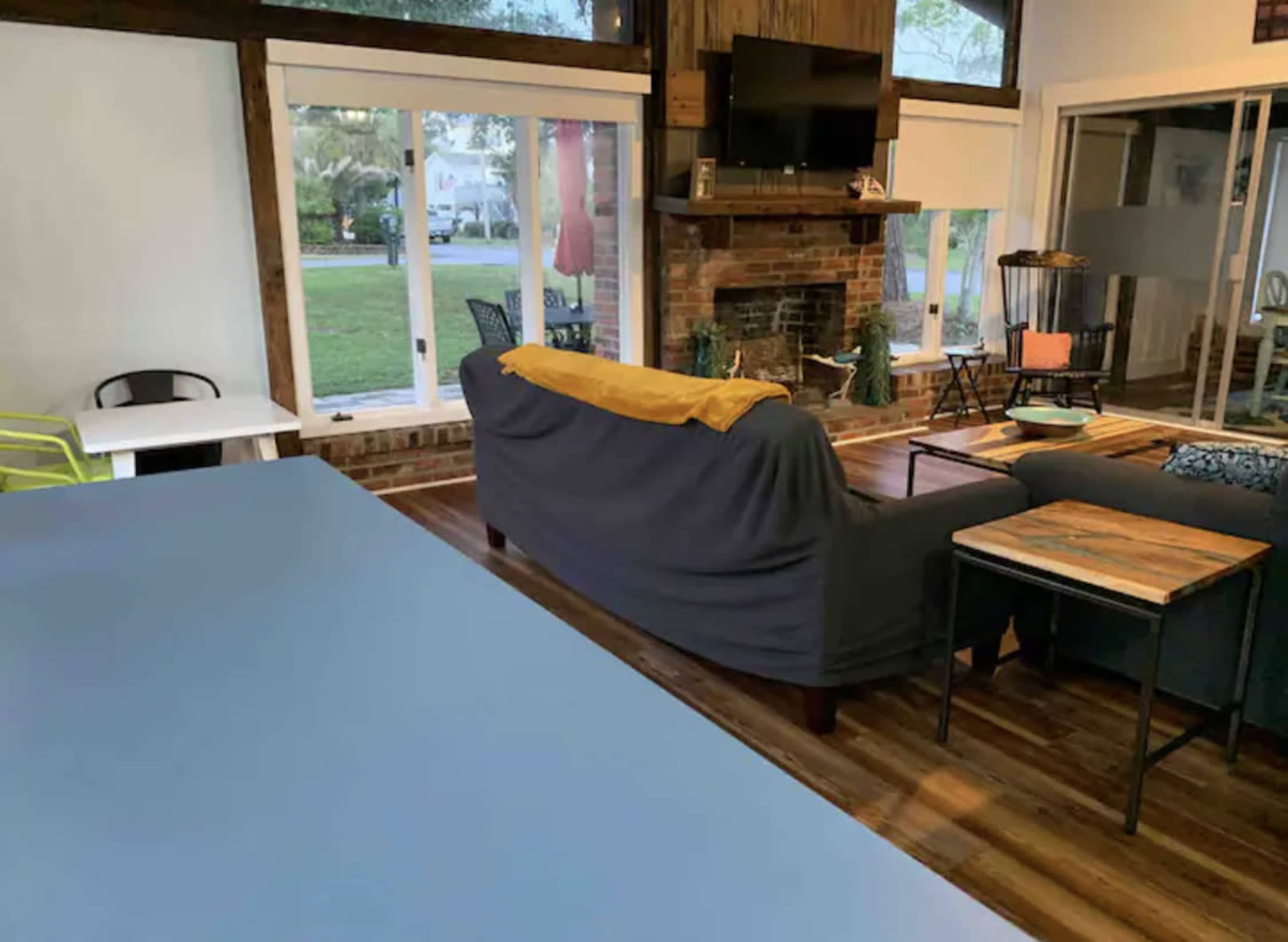The image shows a living room with a brick fireplace, a large sofa covered with a yellow throw, a coffee table, and large windows overlooking a yard.